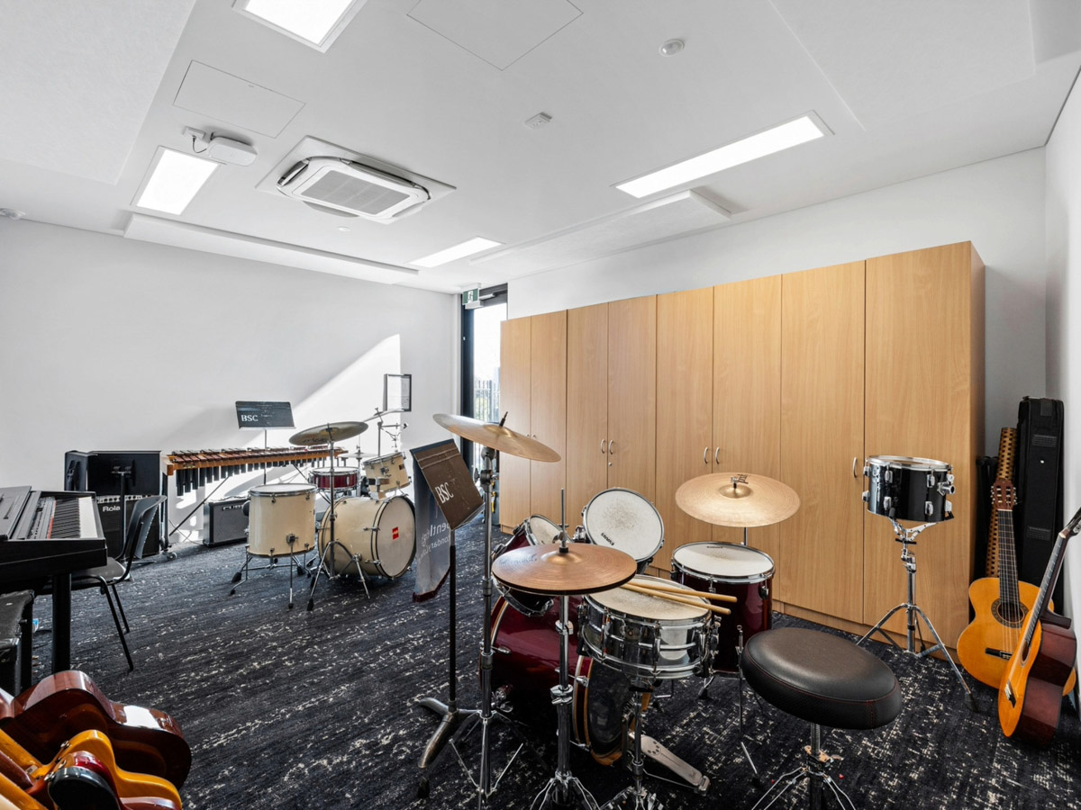 Music classroom with drum kits, guitars, keyboards, and percussion instruments arranged around timber storage cabinets.