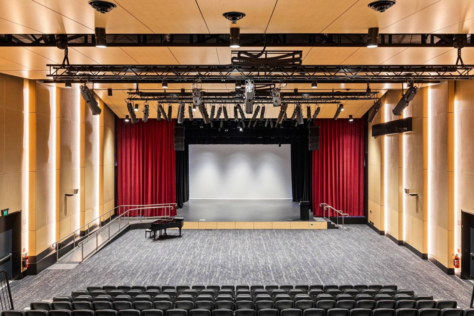 Interior of a modern auditorium with tiered seating, a large stage framed by red curtains, and overhead lighting rigs.