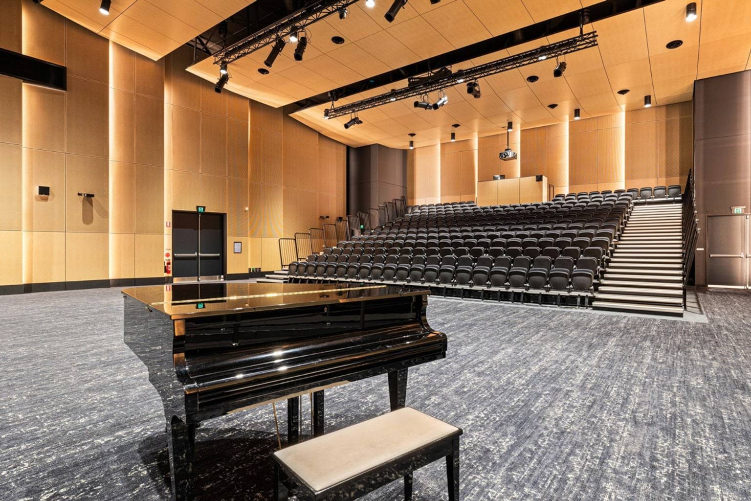 Auditorium interior showing a glossy black grand piano in front of tiered seating and timber-panelled walls.
