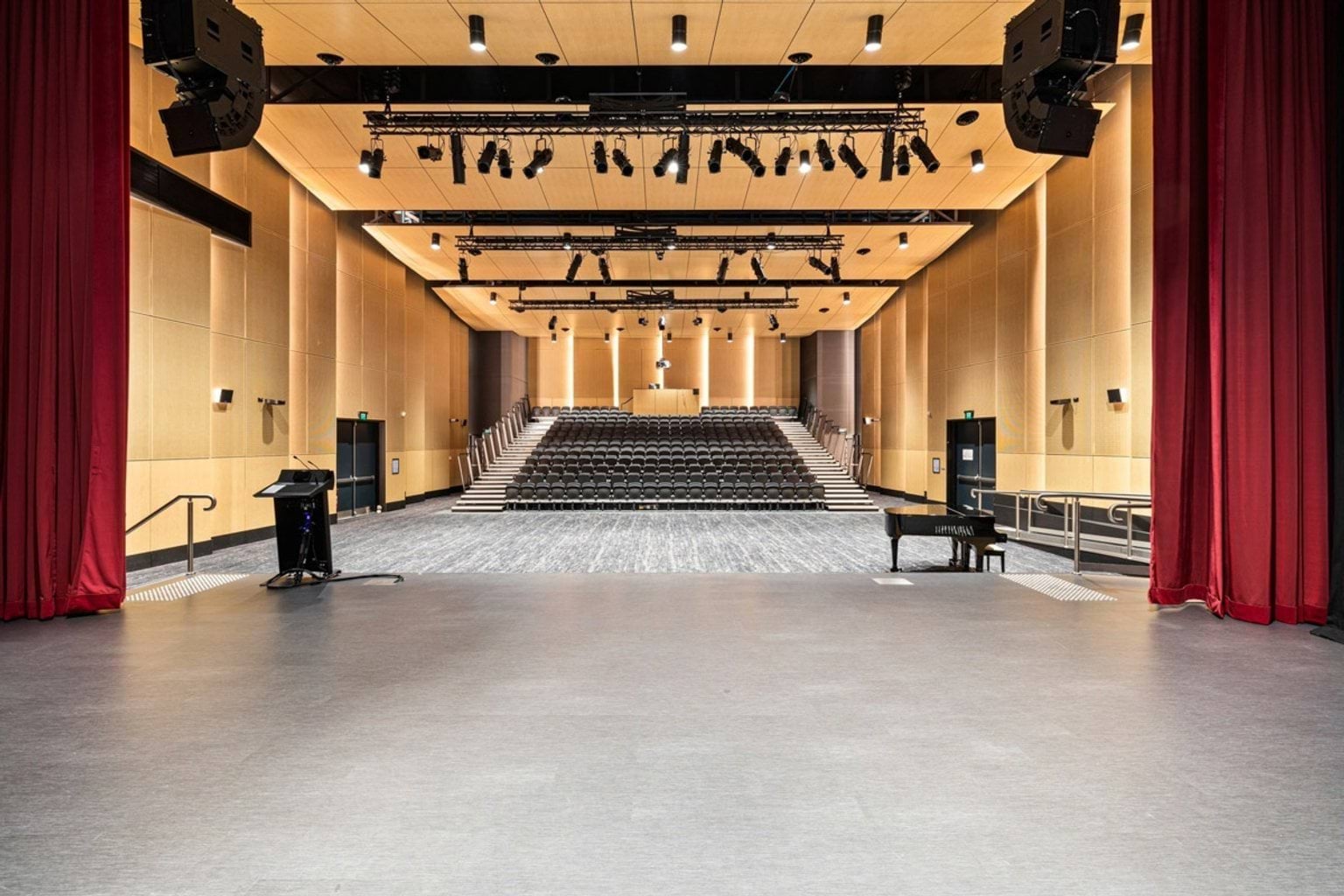 View from the stage of a modern auditorium with tiered seating, timber-panelled walls, and red curtains framing the space.