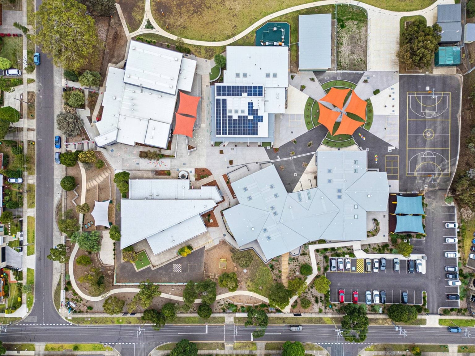 Aerial view of the entire school site showing multiple gabled buildings, orange shade sails, basketball courts, and car parks.