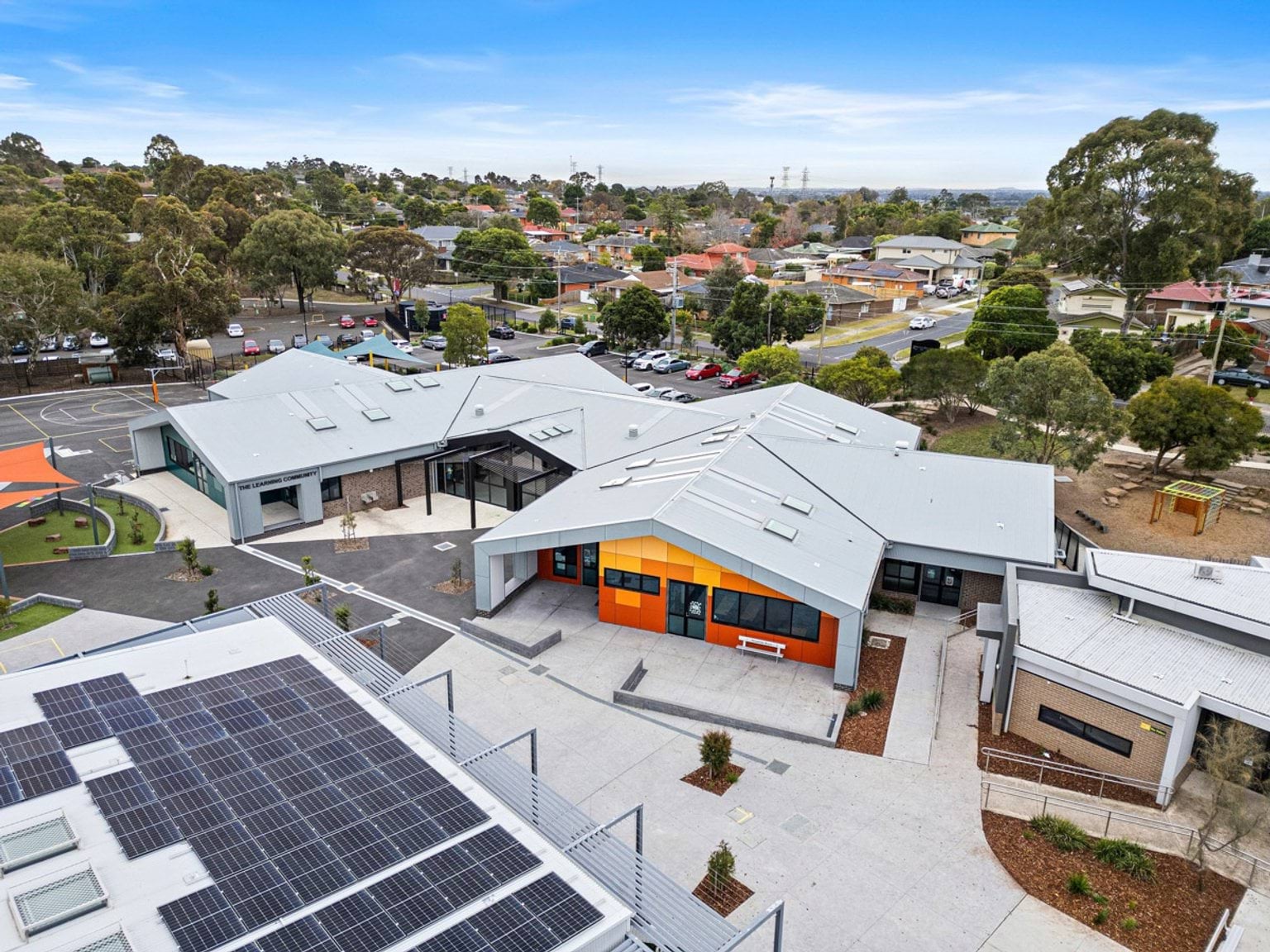 Aerial view of a modern school building with orange cladding and grey roofing, surrounded by paved areas and landscaped garden beds.