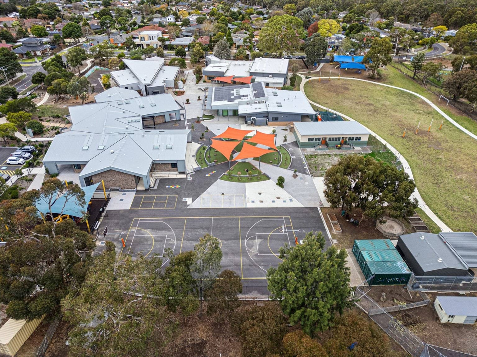 Aerial view of a school campus with multiple gabled buildings, orange shade sails, basketball courts, and surrounding trees and streets.