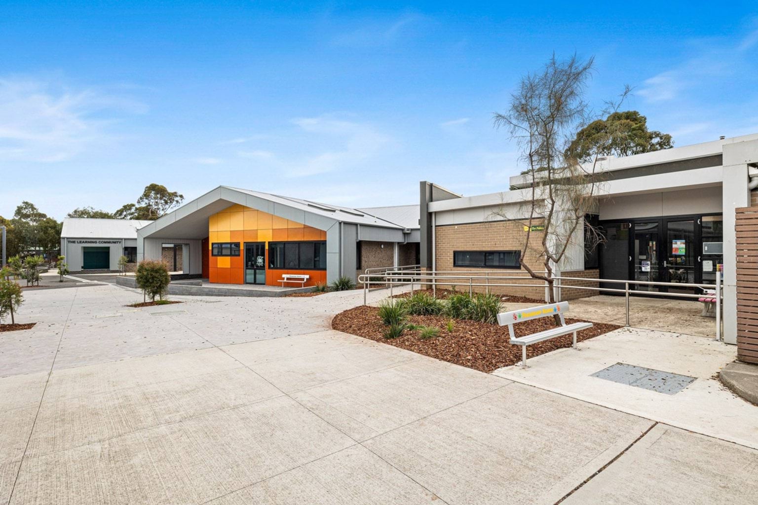 A paved courtyard with landscaped garden beds between modern school buildings. One building has bright orange cladding and a gabled roof.