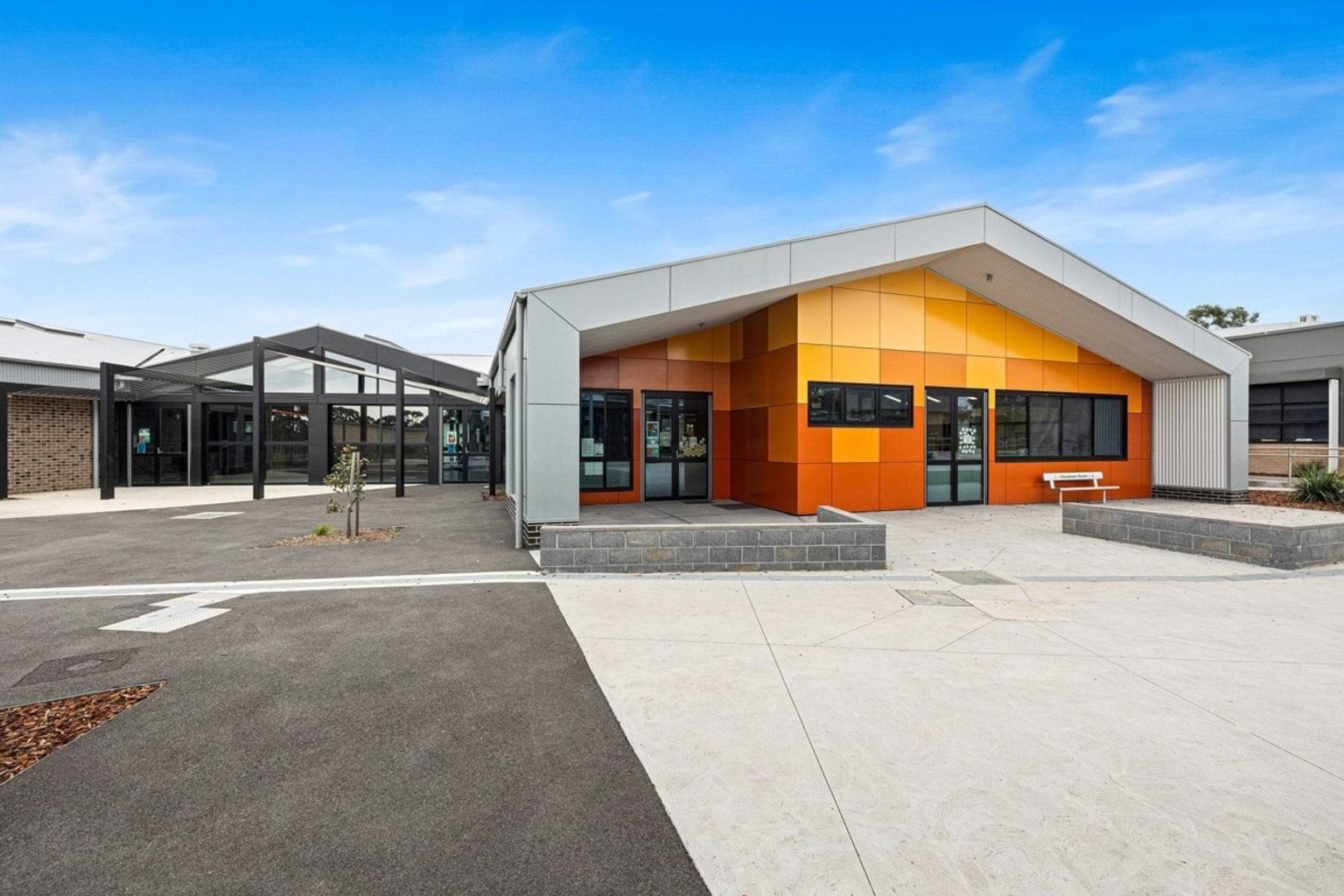 A modern school building with orange walls and a gabled roof, facing a paved courtyard with garden beds and seating.