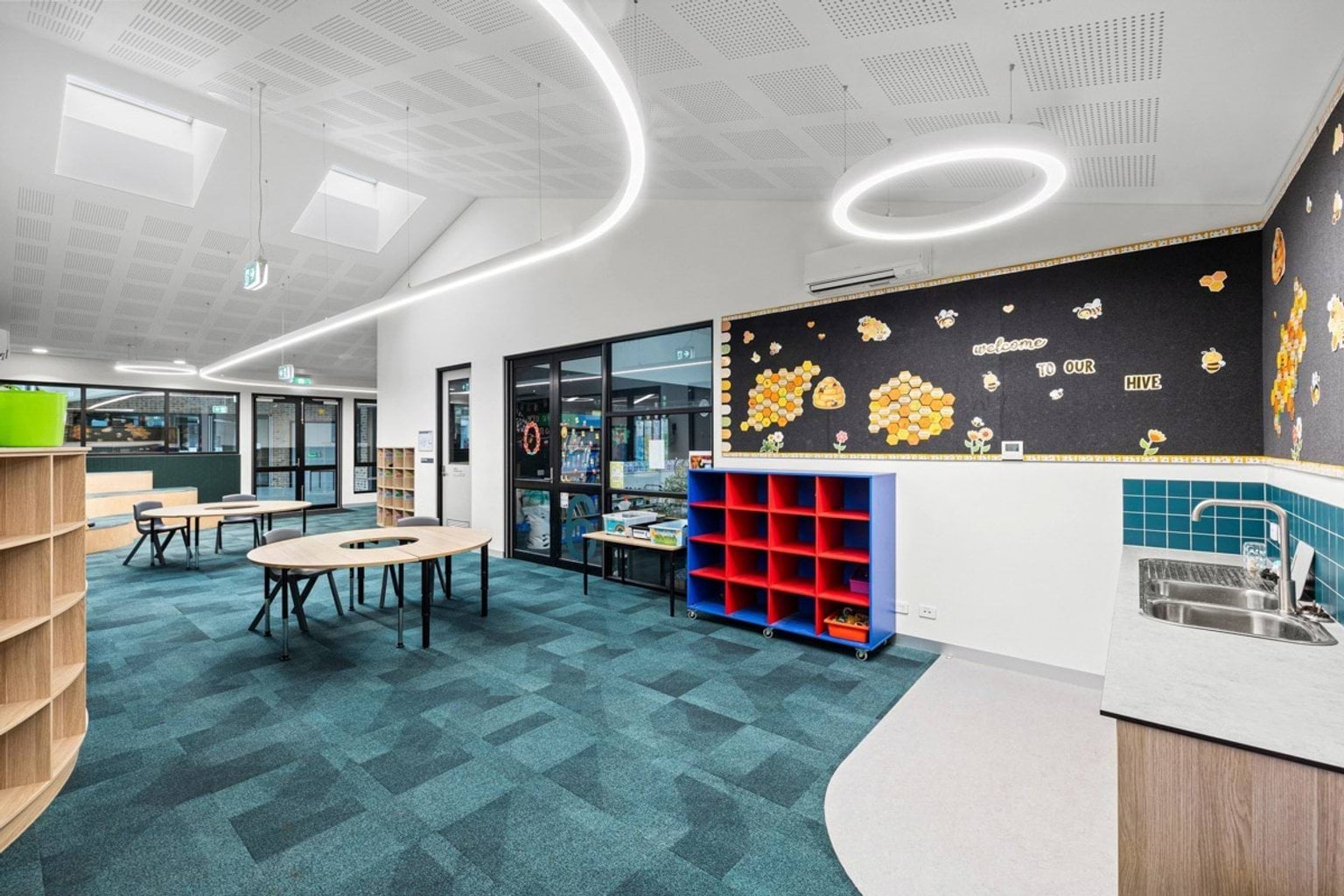 An open learning space with teal carpet, timber tiered seating, round tables, and colourful wall displays under circular ceiling lights.