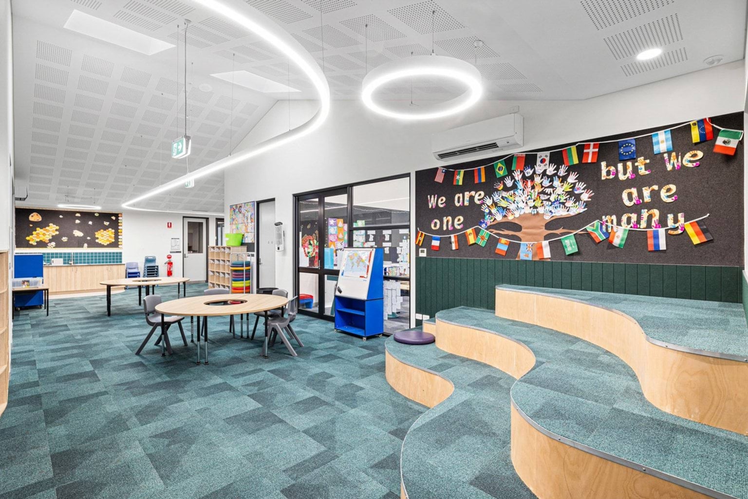 An open learning space with teal carpet, timber tiered seating, round tables, and colourful wall displays under circular ceiling lights.