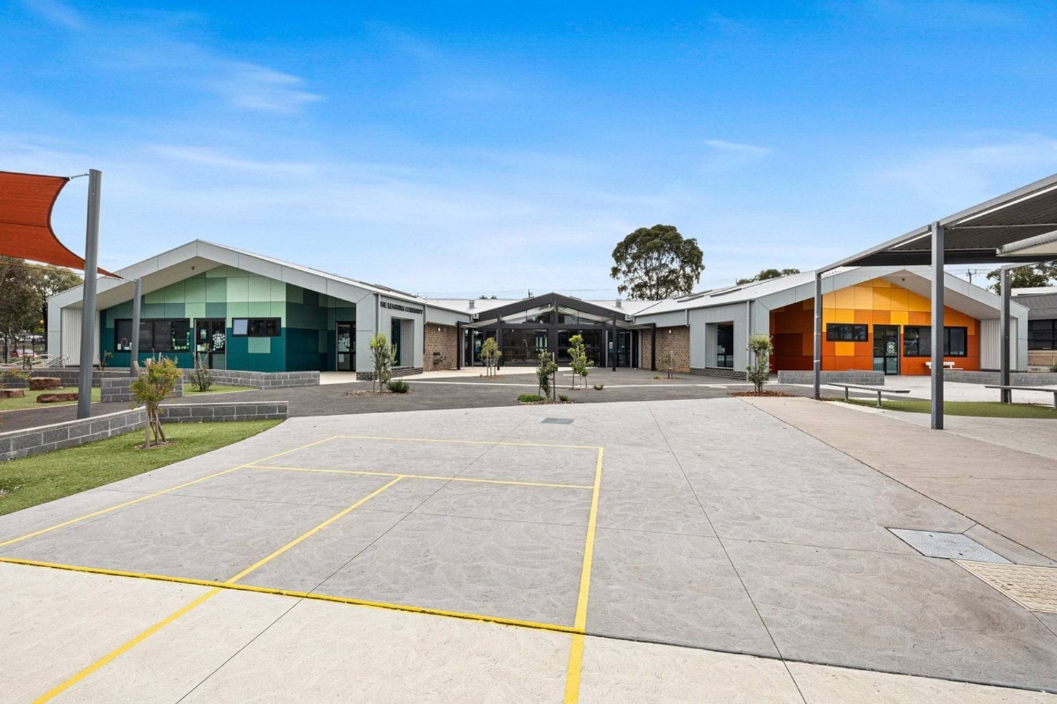 A paved courtyard with garden beds and shade structures between two modern school buildings with green and orange cladding.