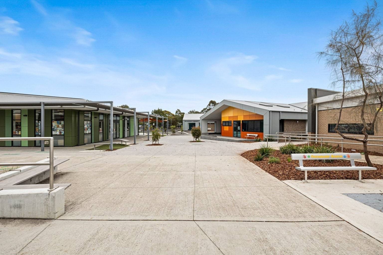 A covered walkway with steel posts and shade panels leading to a modern school building with bright orange cladding.
