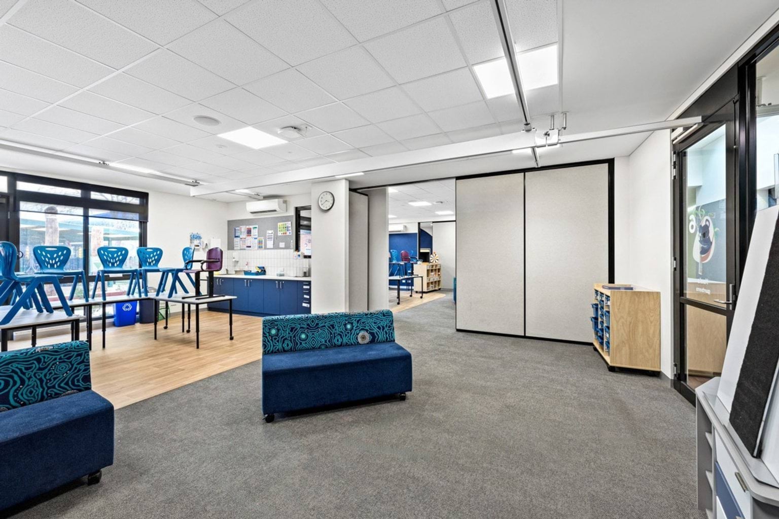 An open learning space with grey carpet, blue chairs stacked on tables, and a kitchenette with blue cabinetry. Movable partitions separate adjoining rooms.