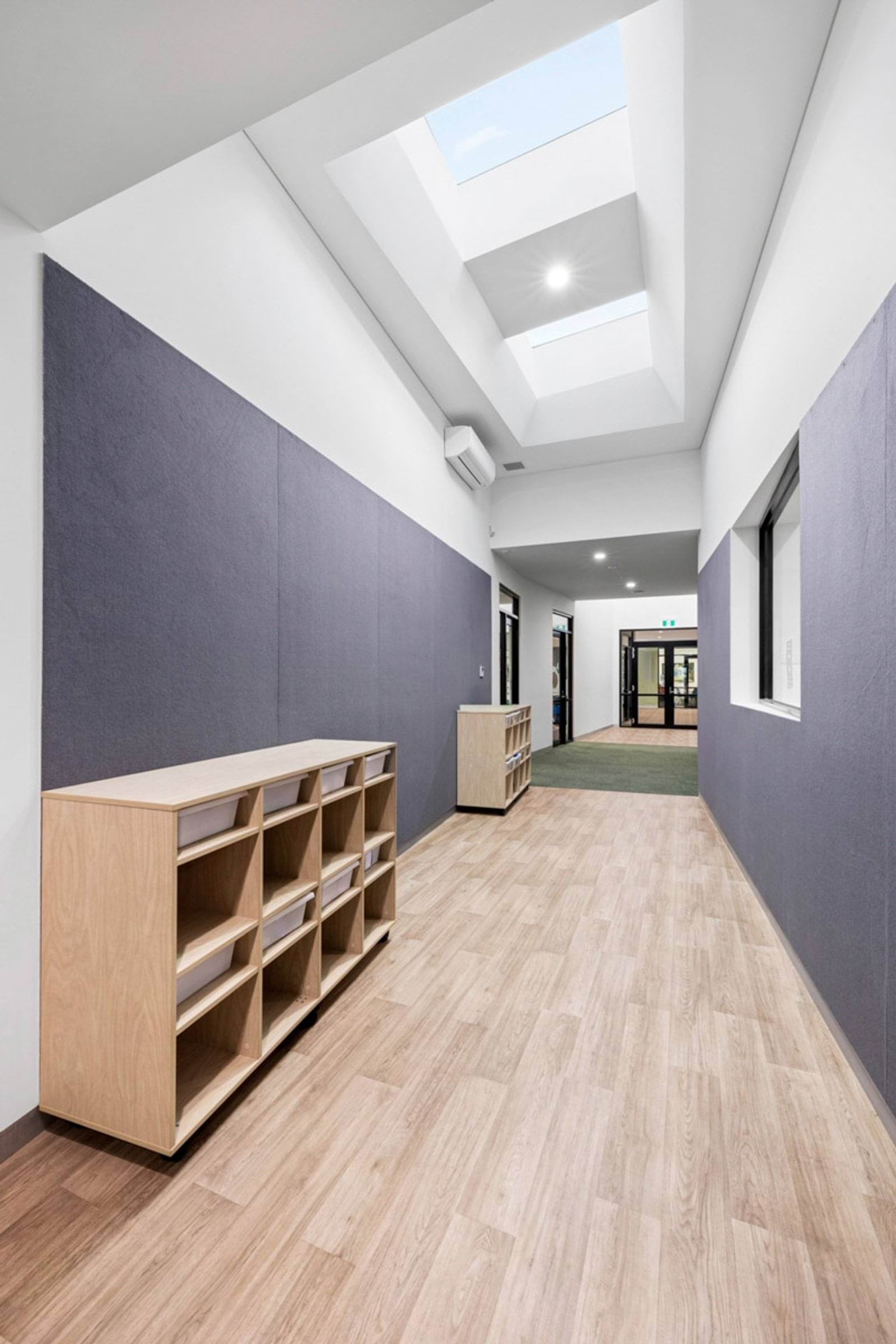 A bright hallway with timber flooring, skylights, and purple wall panels. Timber shelving units are placed along the corridor.