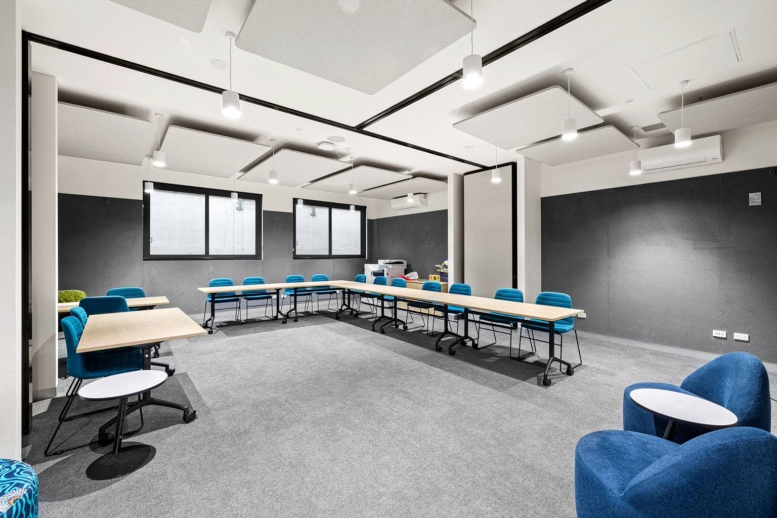 A meeting room with grey carpet, blue chairs arranged around timber tables, and acoustic panels on the ceiling.