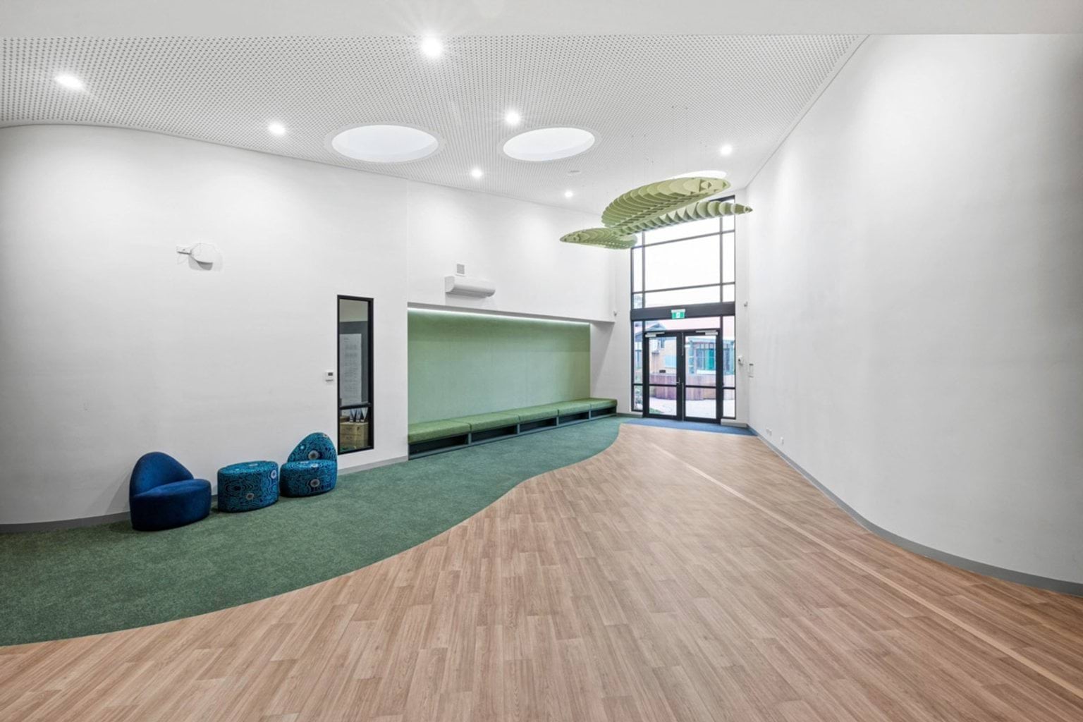 A breakout space with timber flooring, green cushioned bench seating, and circular ceiling lights. Large glass doors lead outside.