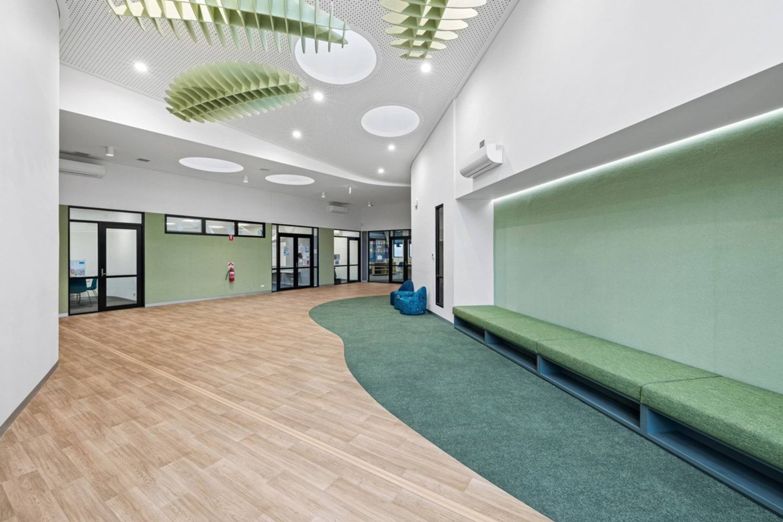 A breakout space with timber flooring, green cushioned bench seating, and circular ceiling lights. Large glass doors open to other areas.