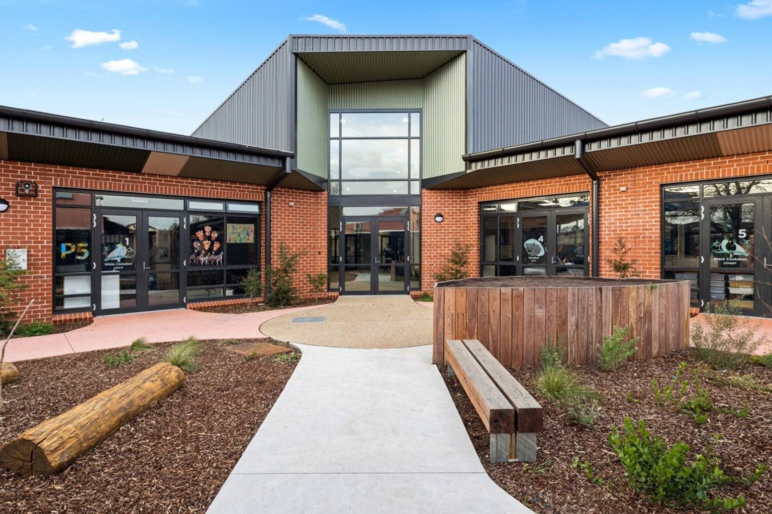 The exterior of a school building with red brick walls, large glass doors, and a landscaped courtyard featuring timber seating and garden beds.