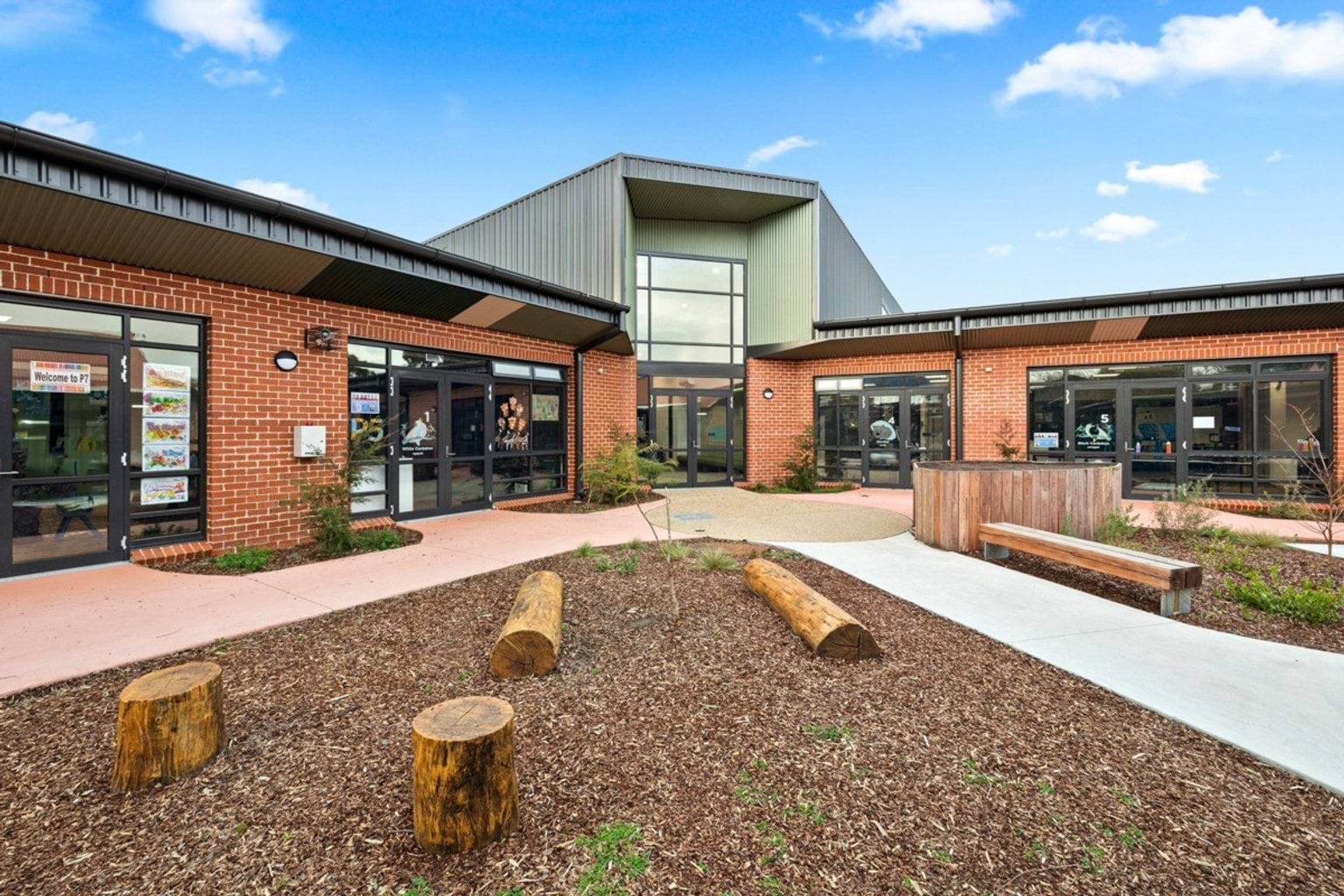 A landscaped school entrance with timber bench seating, garden beds, and log play elements in front of a red brick building with glass doors.