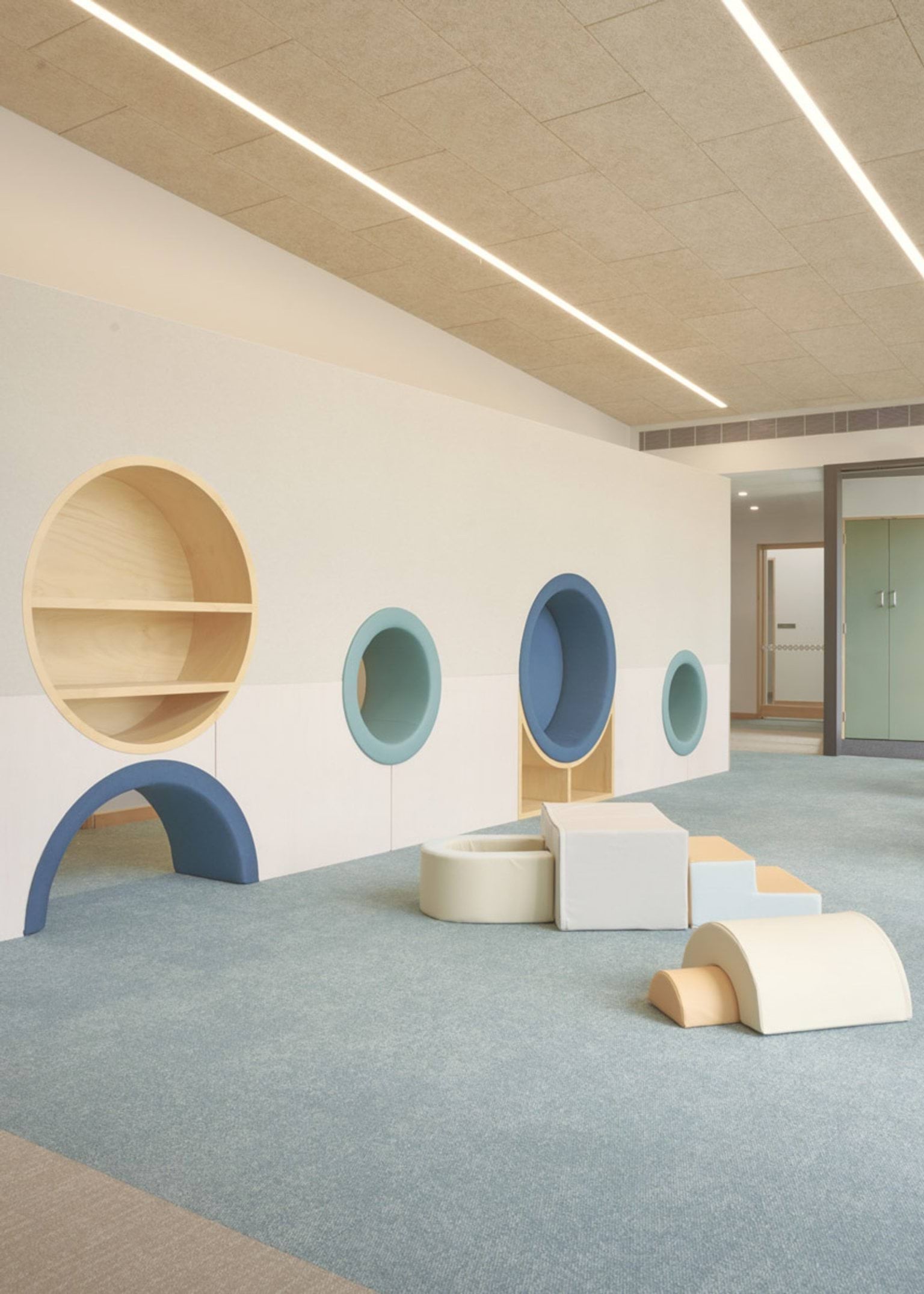 A playroom inside the kindergarten with circular wall cut-outs in shades of blue and timber, and soft foam blocks on the floor. The ceiling has long strip lights and the carpet is pale blue.