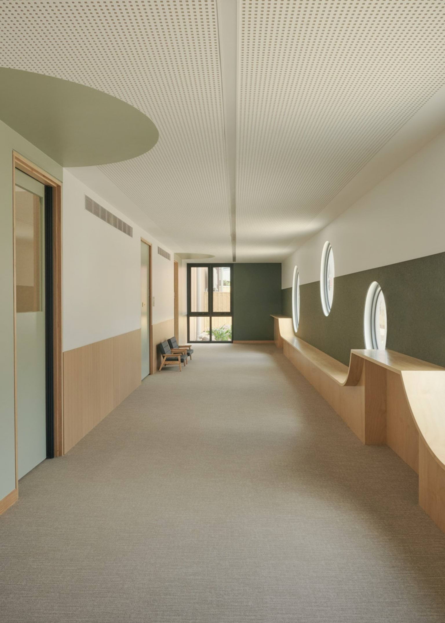 A hallway inside the kindergarten with round windows set into a green wall and timber bench seating below. The ceiling is white with perforated panels and the floor is carpeted in a neutral tone.