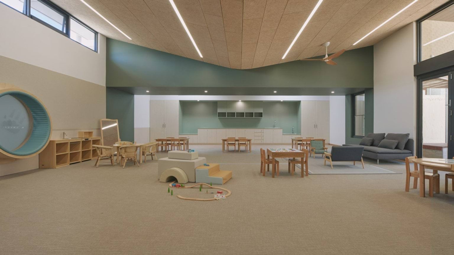 A spacious kindergarten classroom with timber tables and chairs, a grey couch and play equipment on the floor. The back wall has built-in cabinetry and sinks, and the ceiling features angled timber panels with strip lighting.