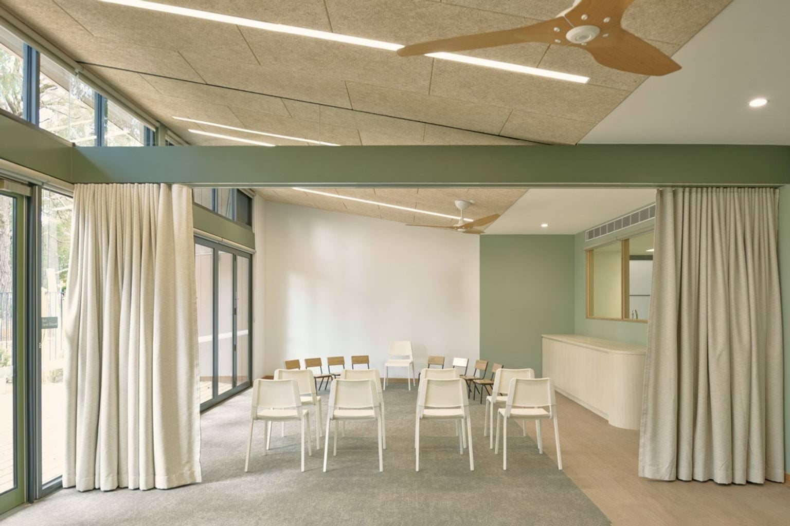 A meeting or group room inside the kindergarten with white chairs arranged in rows facing a green wall. Large windows with curtains line one side and the ceiling has timber panels and a fan.