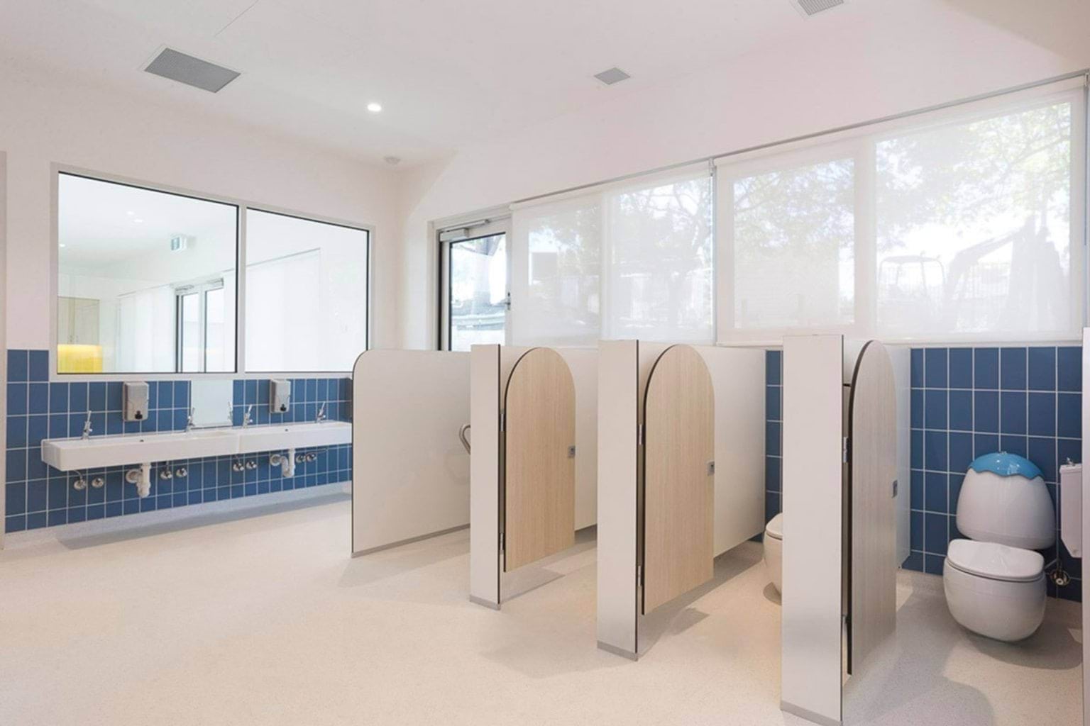 Children’s bathroom with blue tiled walls, cubicles with curved doors and a row of basins with mirrors
