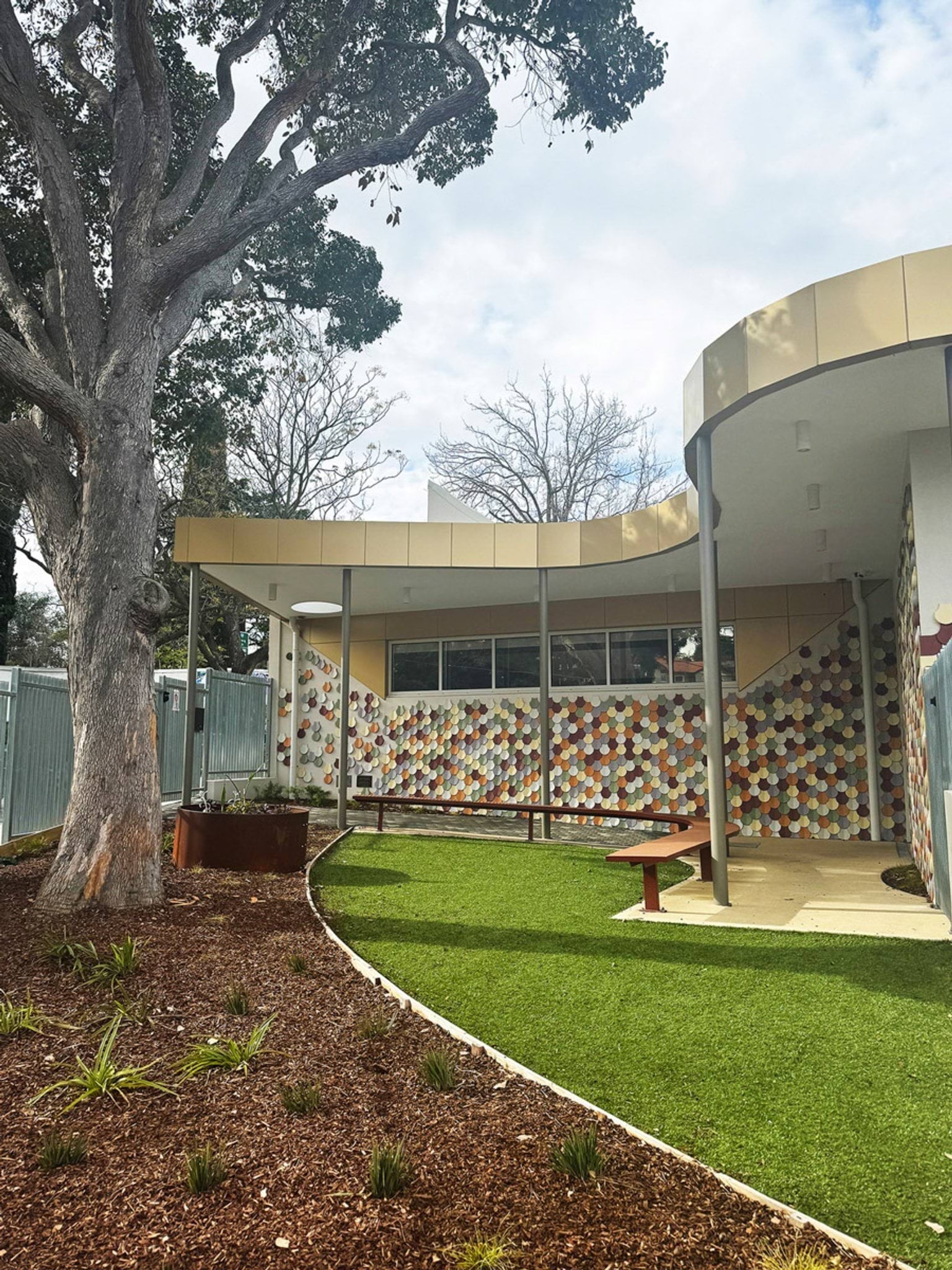 Outdoor play area with grass, curved bench seating and building facade featuring multicoloured tiles