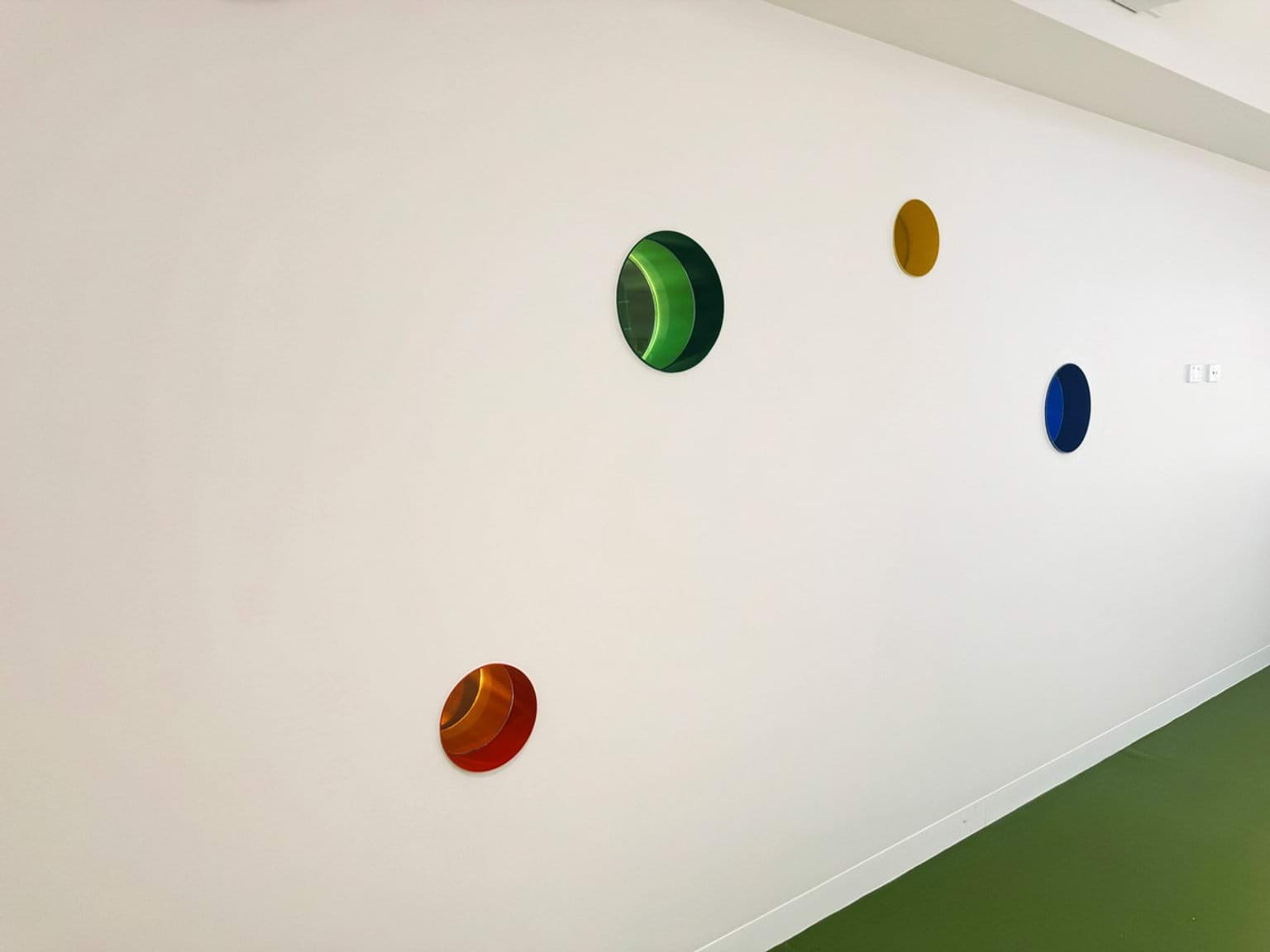White wall with four circular coloured windows in red, green, yellow and blue above a green floor