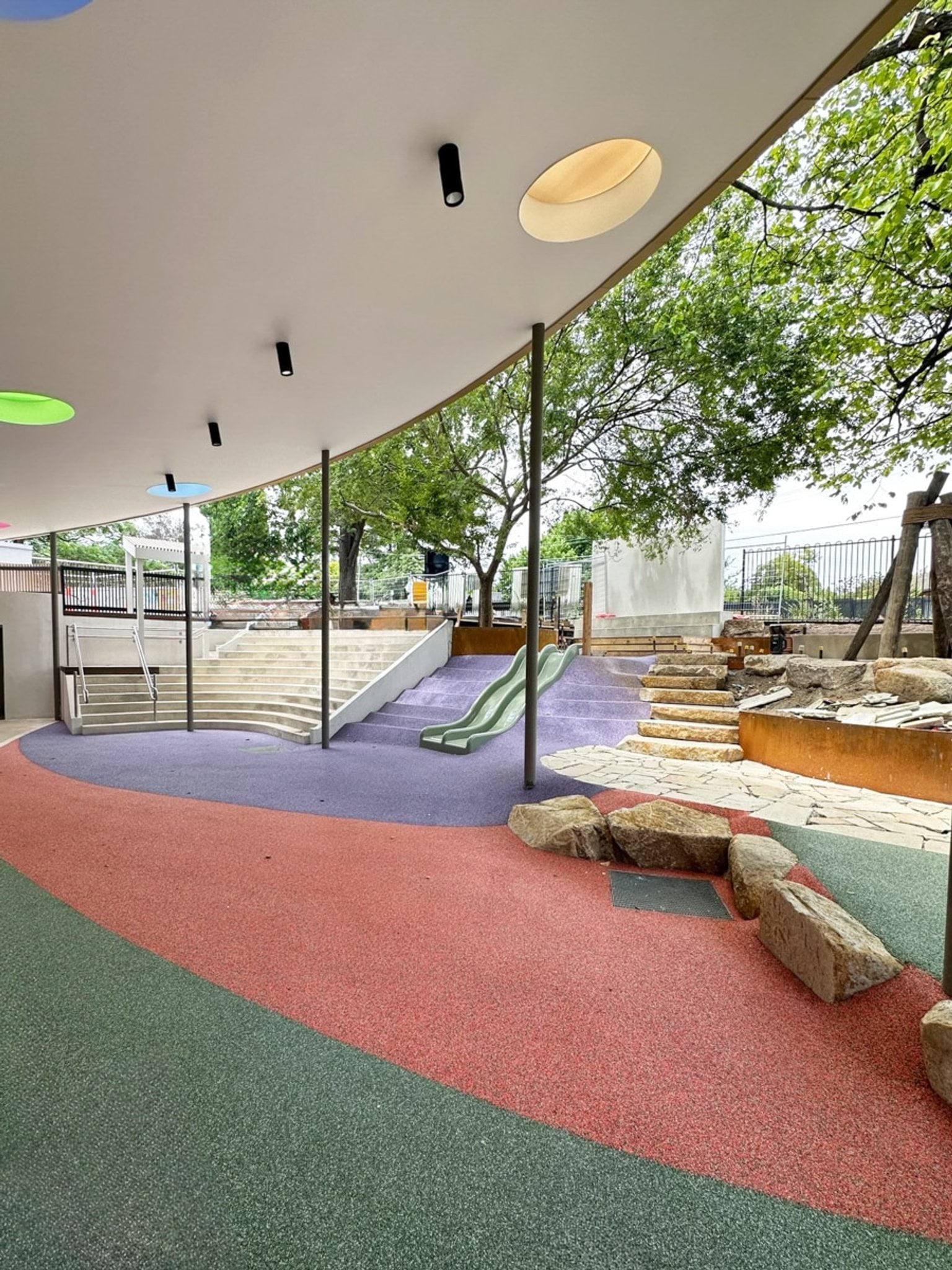 Covered outdoor play area with curved roof, coloured ceiling cut-outs, slides and tiered stone steps