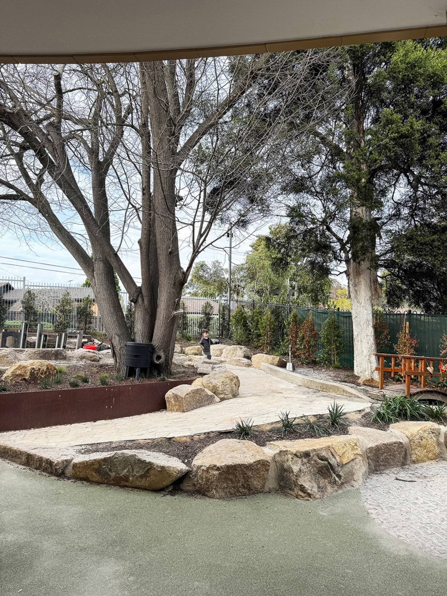 Landscaped outdoor area with stone-edged pathways, large trees and garden beds beside a preschool building