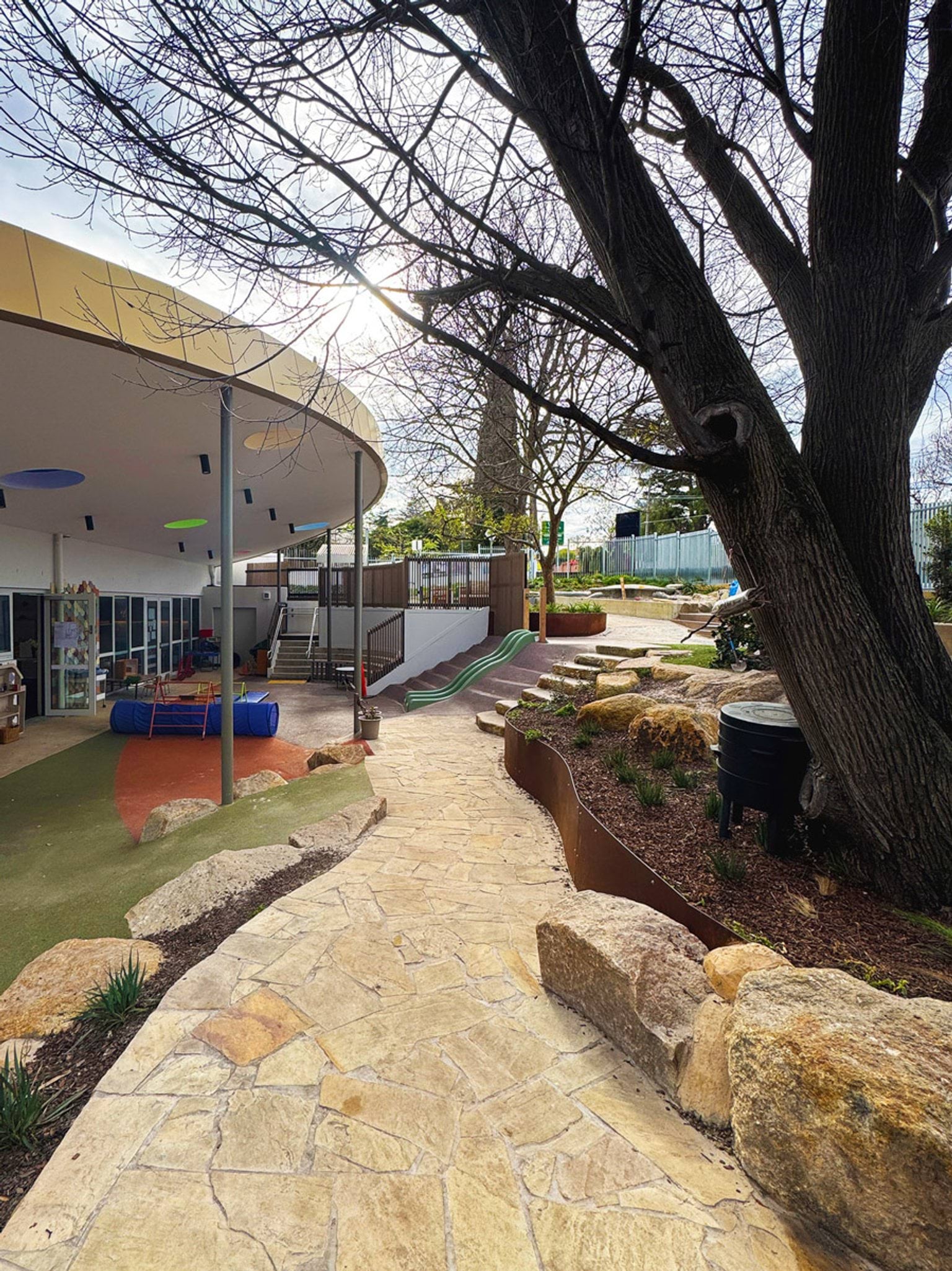 Outdoor play area with curved roof, coloured flooring, slides and stone-edged pathways surrounded by trees
