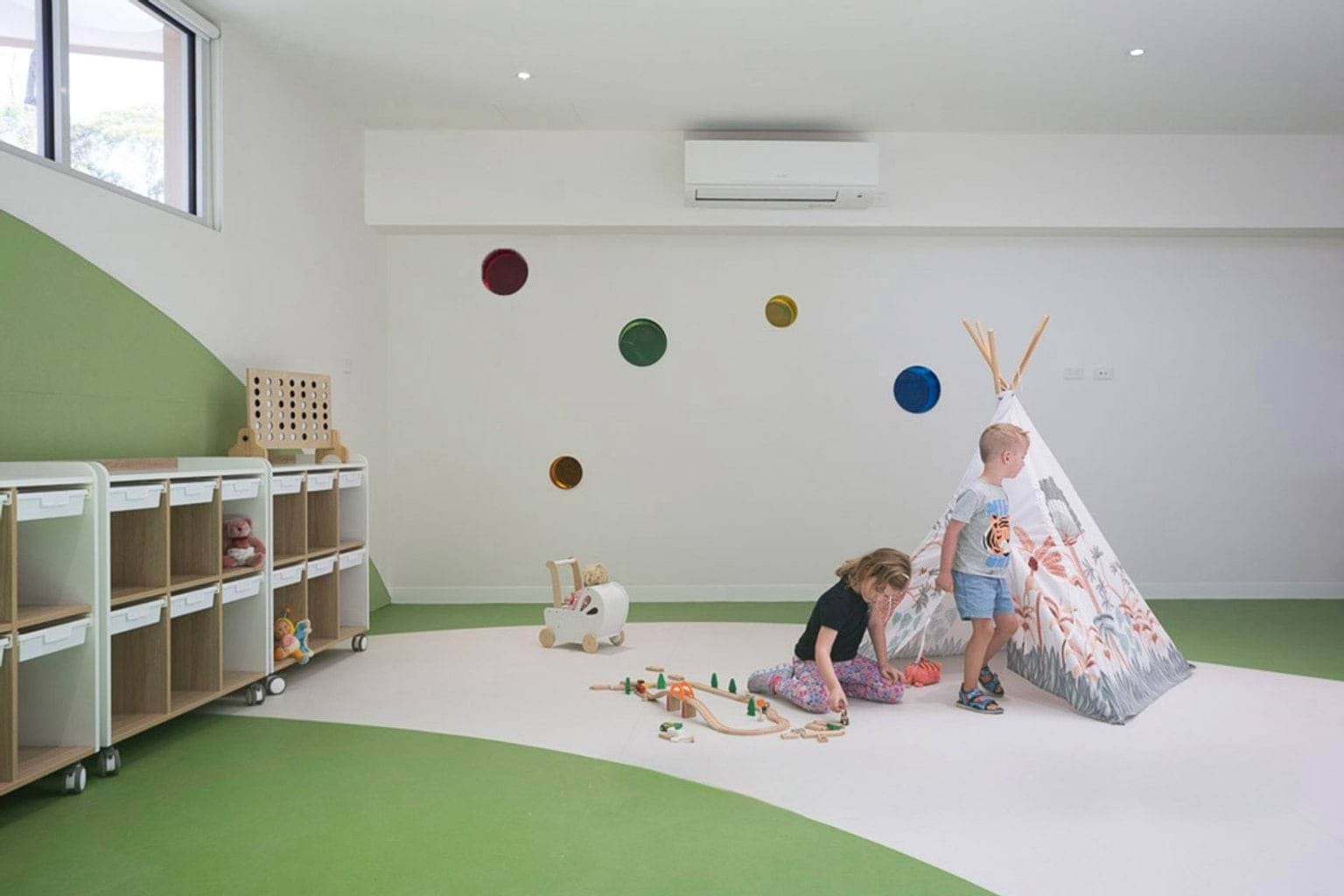 Indoor playroom with green and white flooring, storage units, a teepee and two children playing with toys