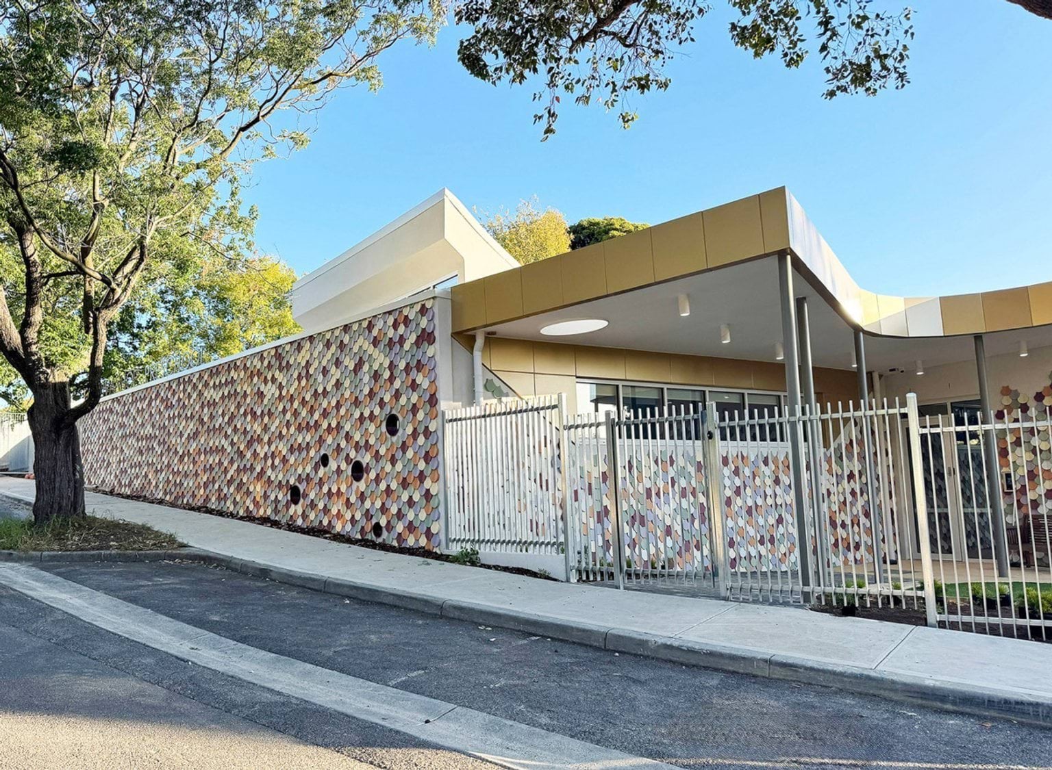Exterior view of preschool building with multicoloured tile facade, curved roof and metal fencing along footpath