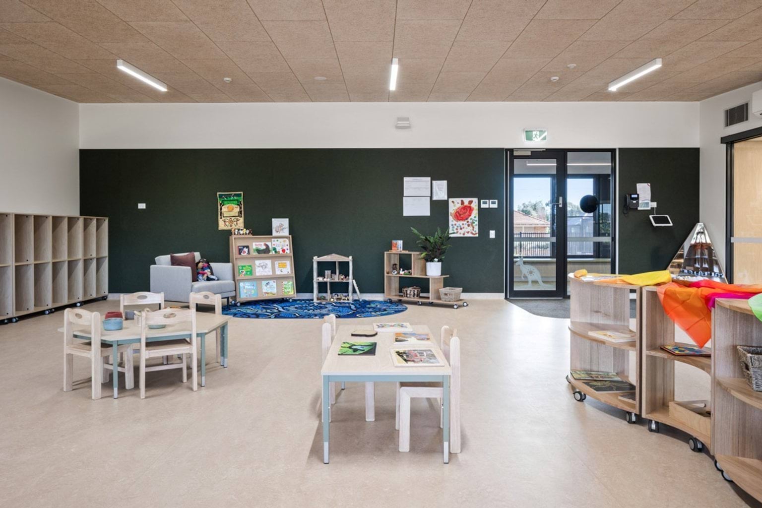 Early learning classroom with small tables and chairs, open shelving, reading corner and educational displays on dark feature wall