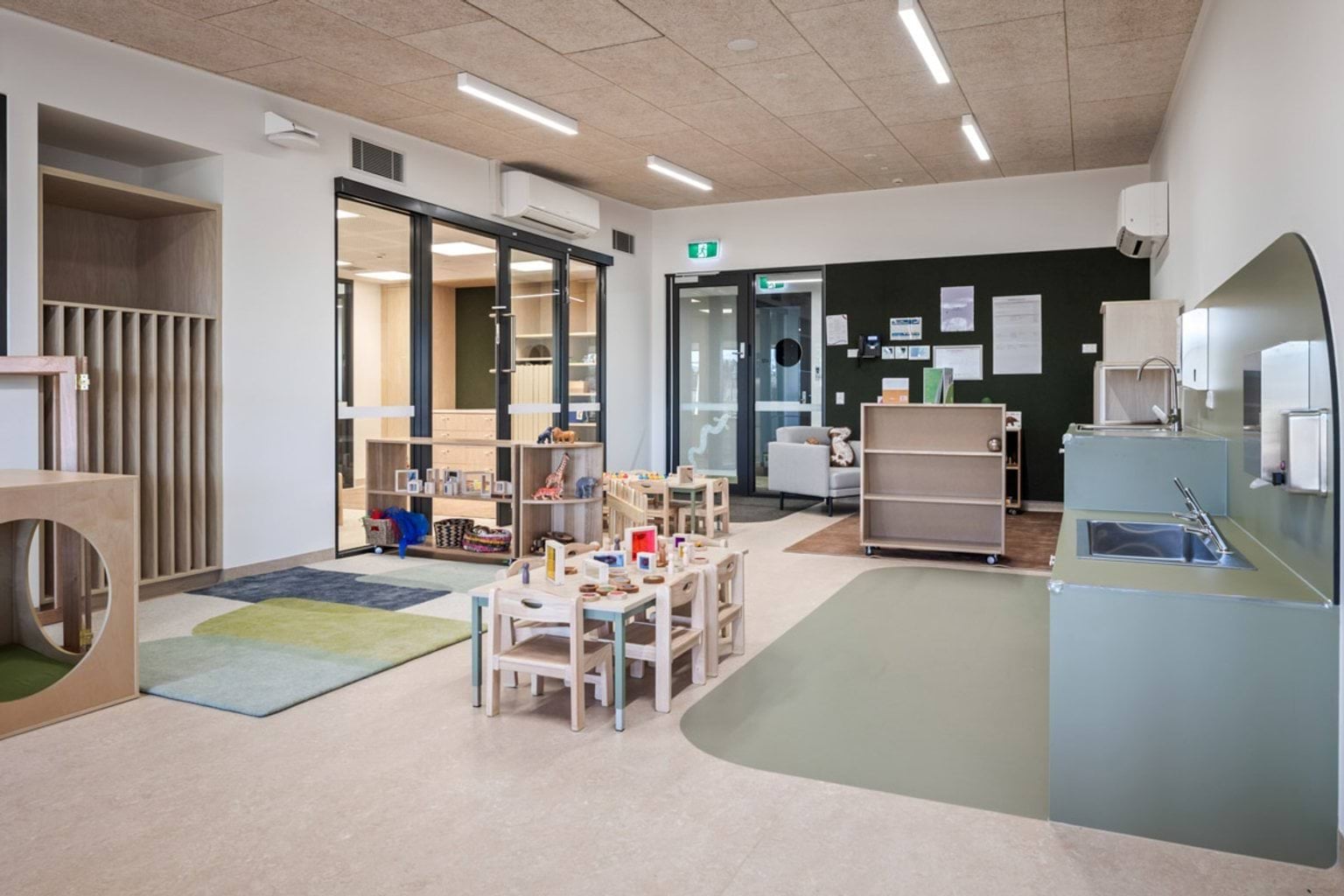 Early learning playroom with activity tables, storage units and play mats, glass partitions opening to adjacent rooms