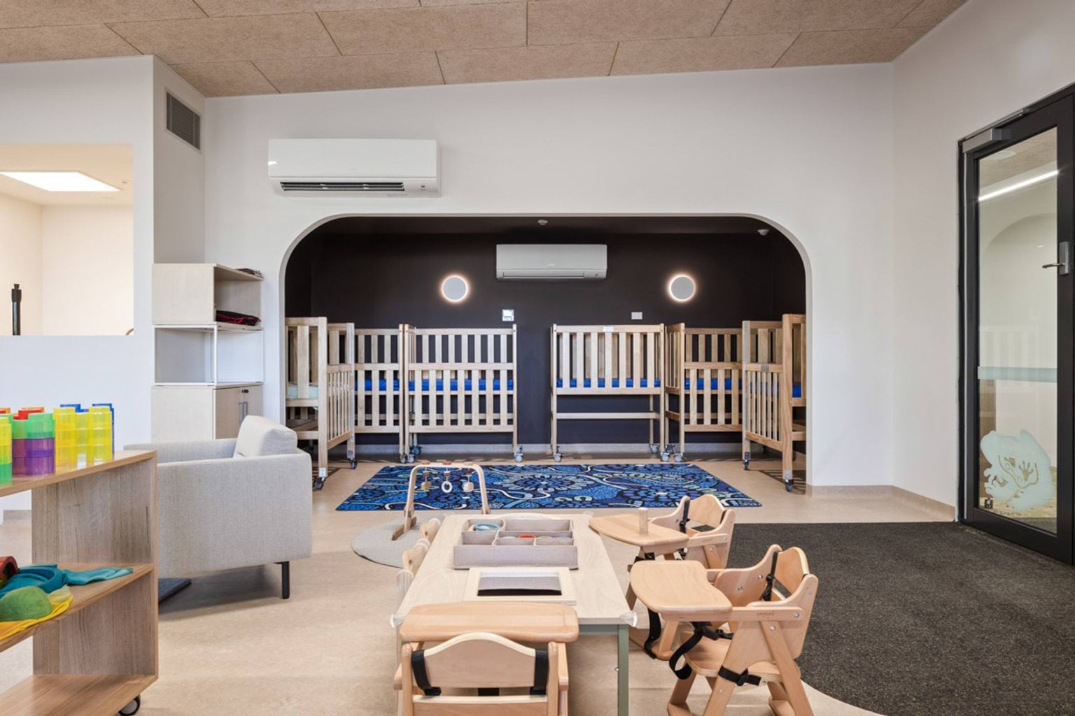 Infant sleep area with timber cots arranged under arched opening, adjoining play space with small tables and chairs
