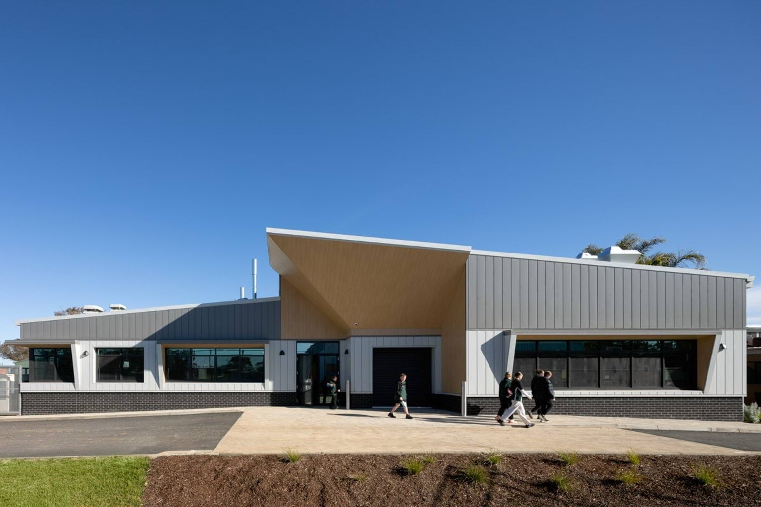The front of the school building with a timber feature above the entrance and grey and white exterior. Students are walking along the paved area in front, and the sky is clear and blue.