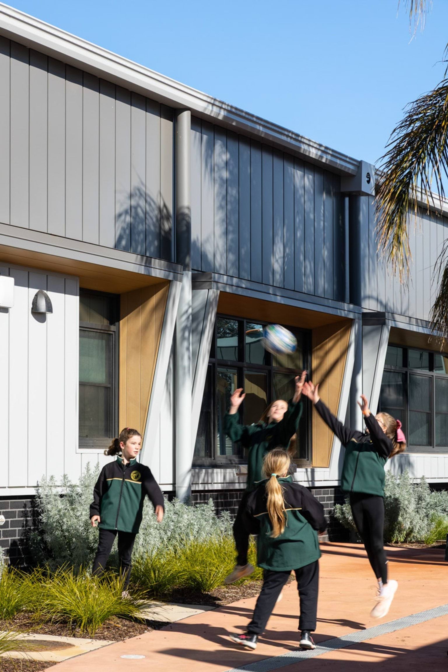 Students playing with a ball in a paved outdoor area next to the school building. The building has a grey and white exterior with timber accents around the windows, and there are garden beds with greenery.