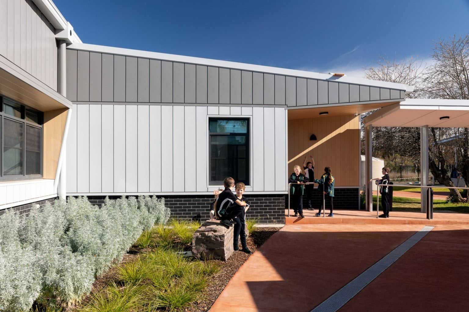 A landscaped courtyard outside the school building, with garden beds and a paved area. Students are sitting on a rock and standing near the entrance, which has timber panels and a covered walkway.