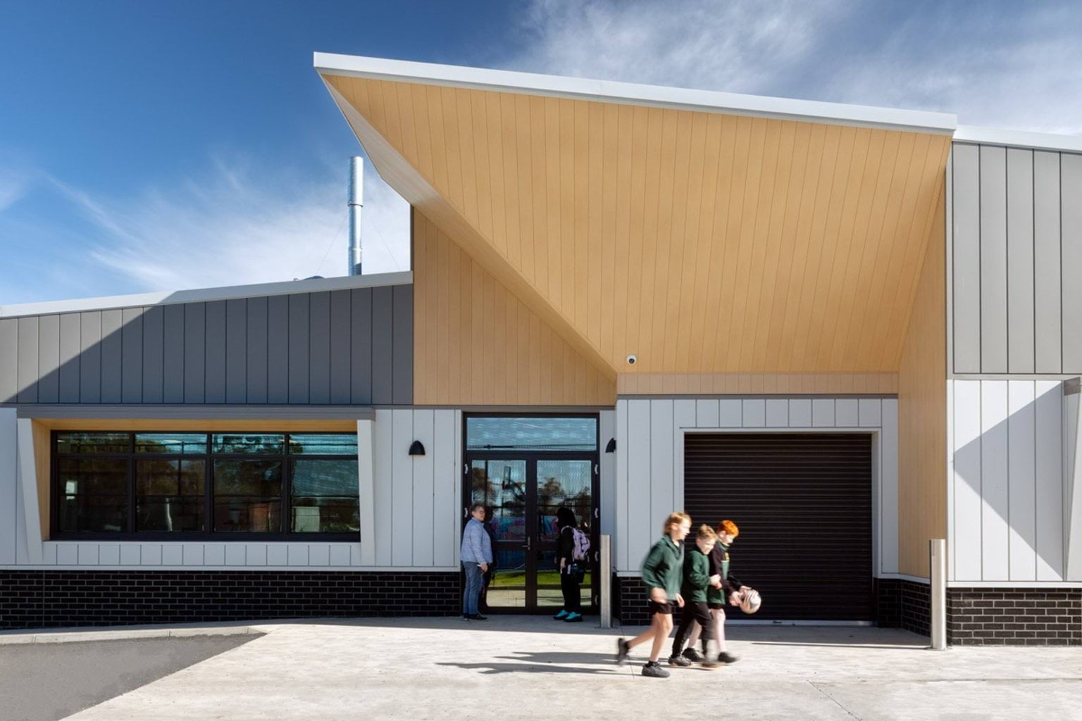 Exterior of the school building, featuring angled timber panels above the entrance and grey and white cladding. A group of students is walking past the entrance, and the sky is clear and blue.