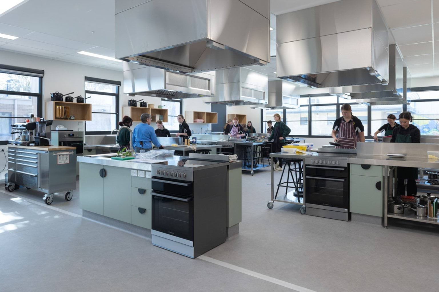 A modern kitchen classroom with stainless steel benches, ovens, and large overhead range hoods. Students are preparing food at different stations, and the room has big windows letting in natural light.