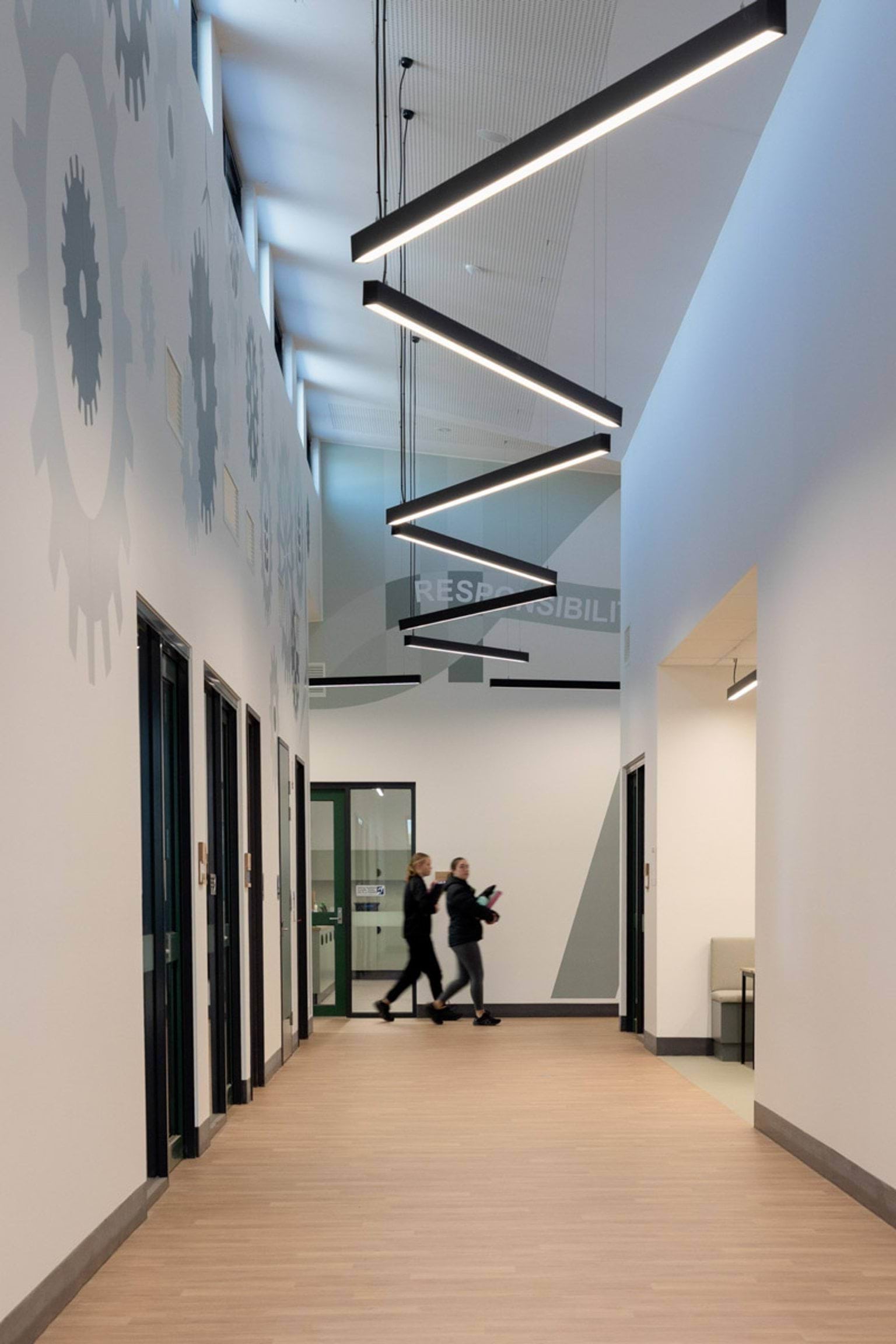 A wide hallway inside the school with light timber flooring and geometric pendant lights hanging from the ceiling. The walls have gear-shaped graphics, and two students are walking through the space.