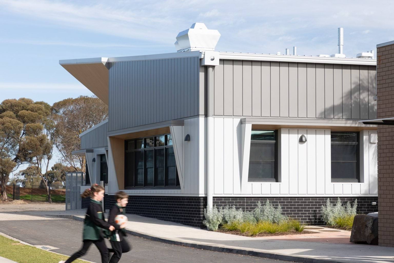 The side of the school building with grey and white exterior and black brickwork at the base. Two students are walking along the path in front, and there are garden beds with shrubs beside the building.