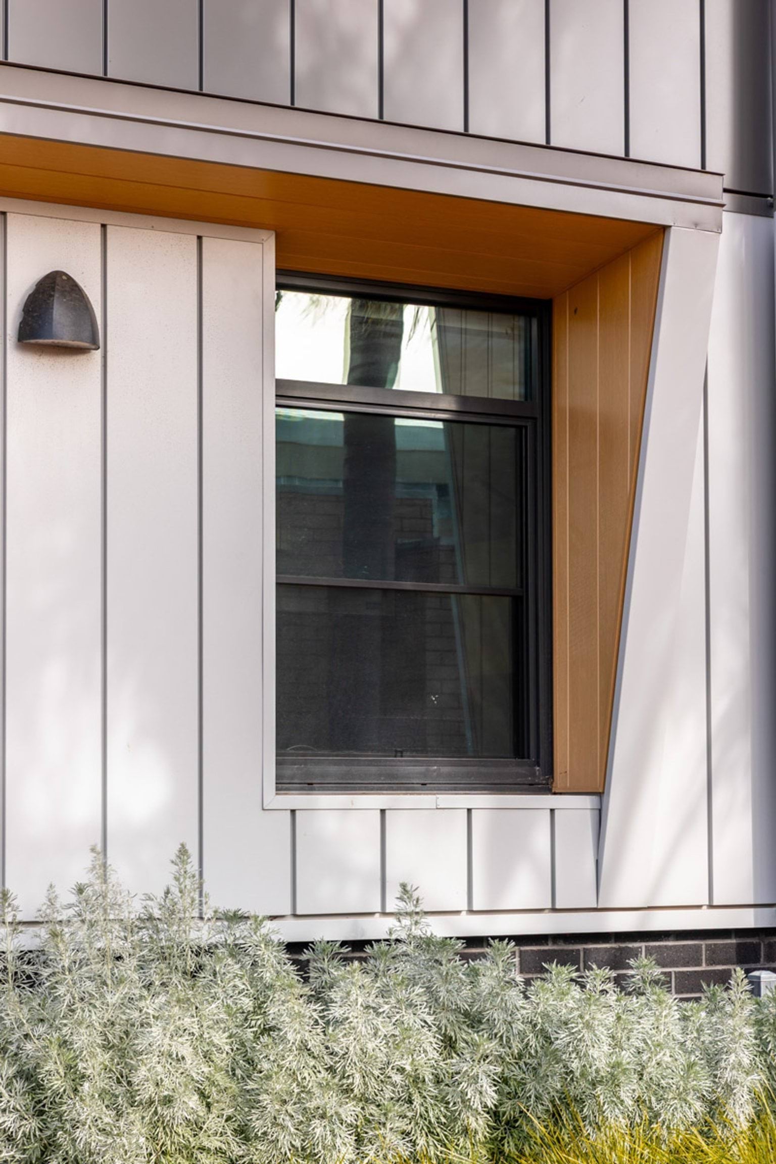 A close-up of a school building window framed by angled timber panels. The wall has grey and white walls, and there are shrubs planted along the base.
