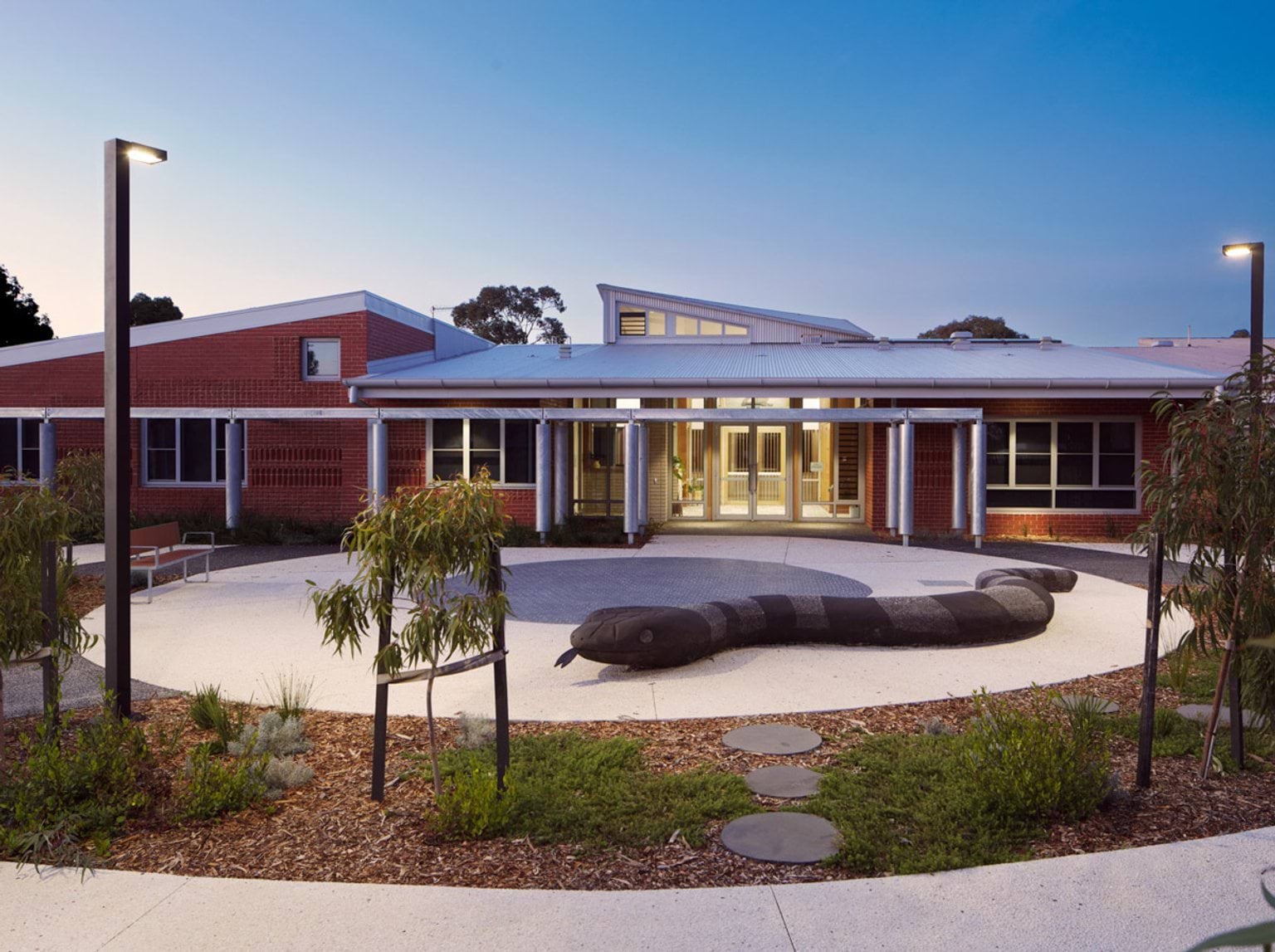 The exterior of the school with red brick walls and a white roof. The courtyard has curved paths, garden beds, and a large snake sculpture in the centre.