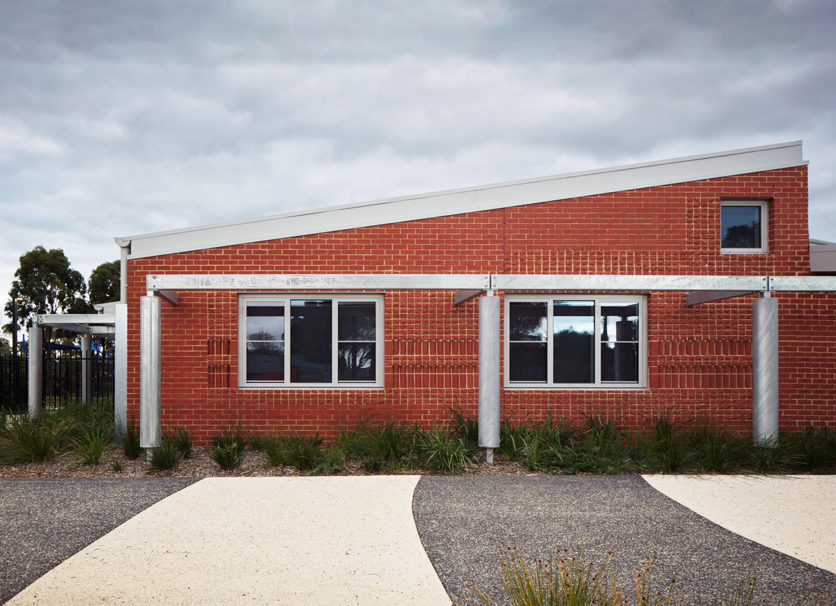 The side of the school building with red brick walls, silver columns, and rectangular windows. Curved concrete paths lead up to the entrance.