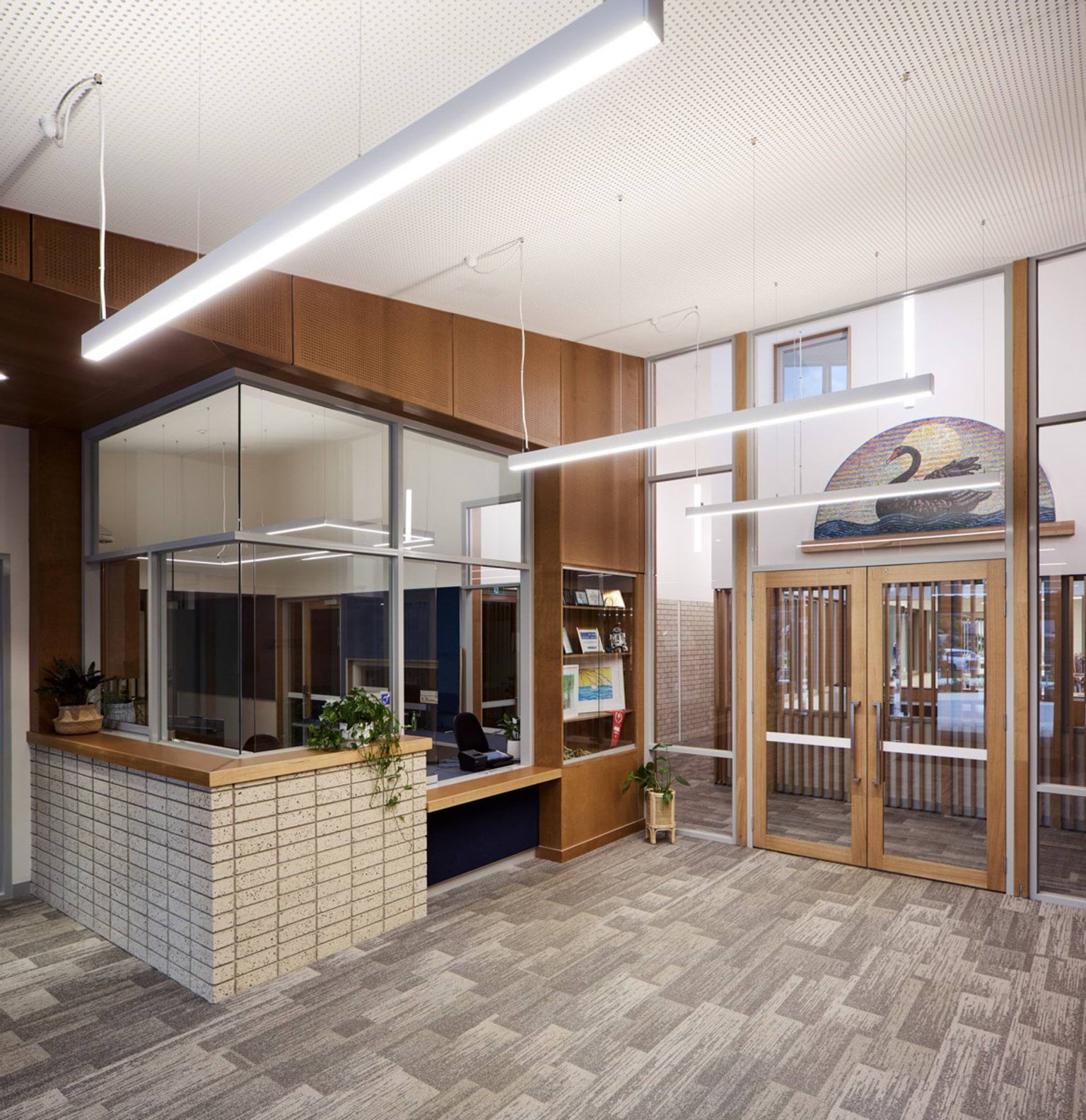 A reception area with a timber-framed glass office and a counter with plants. Pendant lights hang from the ceiling above.