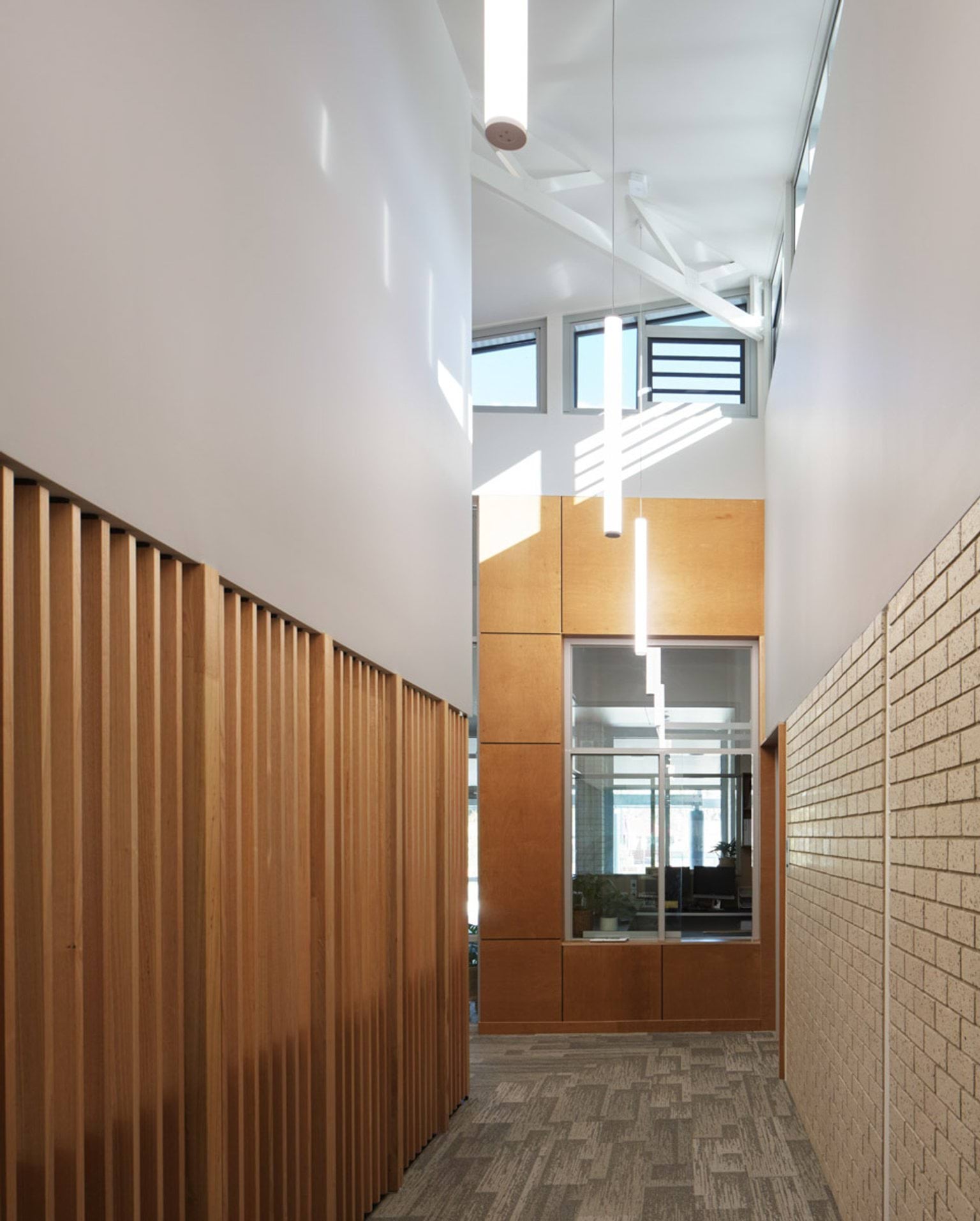 A hallway with timber slats on one side and light brick on the other. High windows and pendant lights brighten the space.