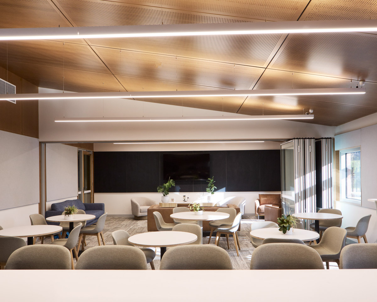 An indoor lounge with round tables and cushioned chairs. The space has a timber ceiling with linear lights and a large screen on the far wall.