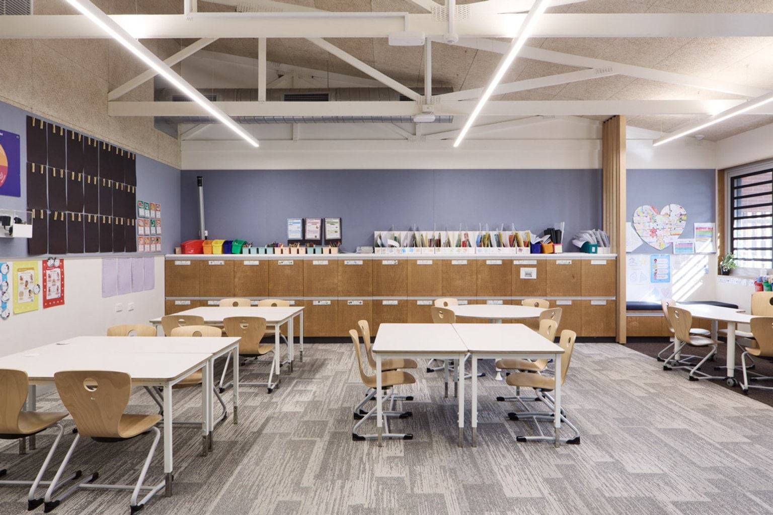 A classroom with white tables, timber chairs, and a wall of storage cupboards. Bright displays and student work decorate the walls.