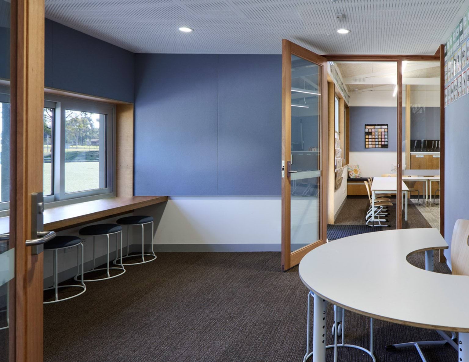 A breakout area with a curved table and stools along a window bench. Glass doors open into a classroom beyond.